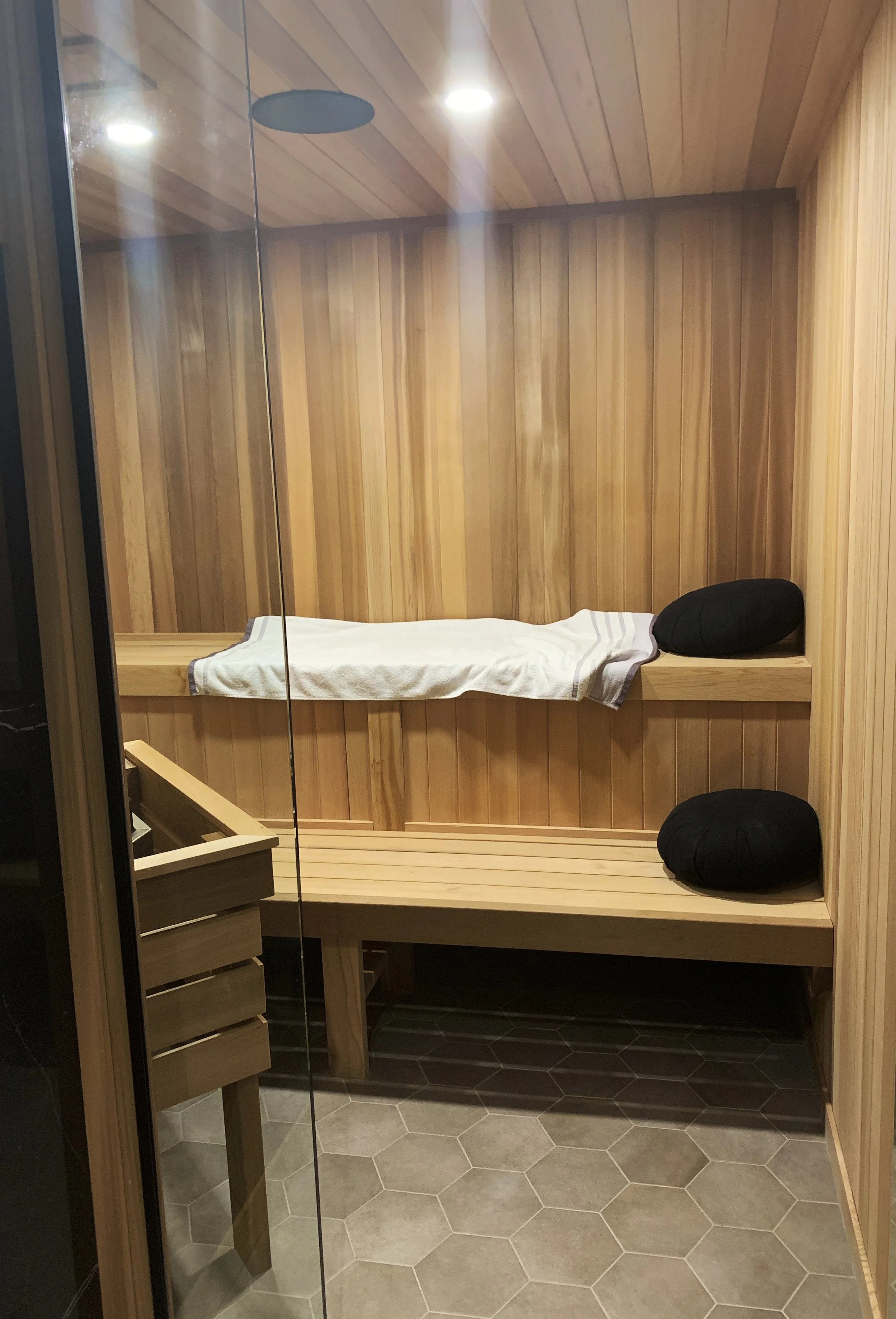 Modern sauna interior with light wood paneling, hex tile floor, glass door, black cushions, and recessed lighting—features cozy bench seating and sleek design ideal for luxury wellness spaces