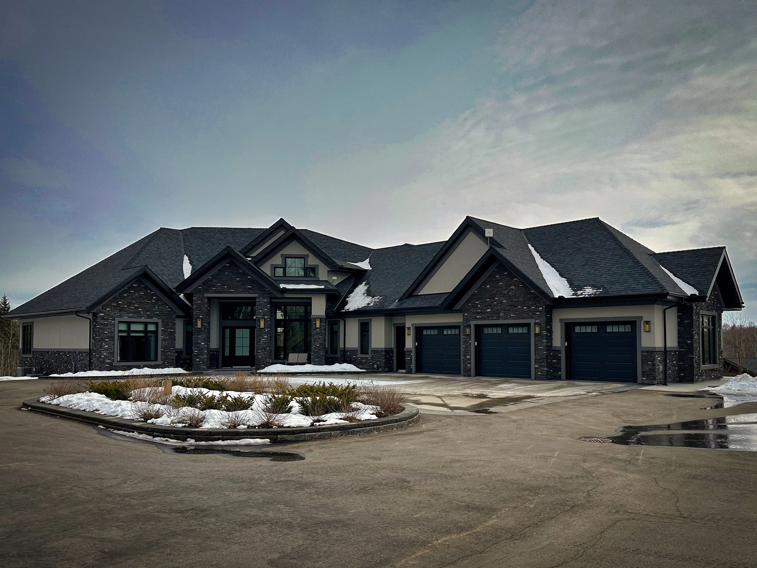 A large modern house with a dark stucco and stone exterior, three garage doors, and snow on the roof, with a mostly overcast sky in the background.