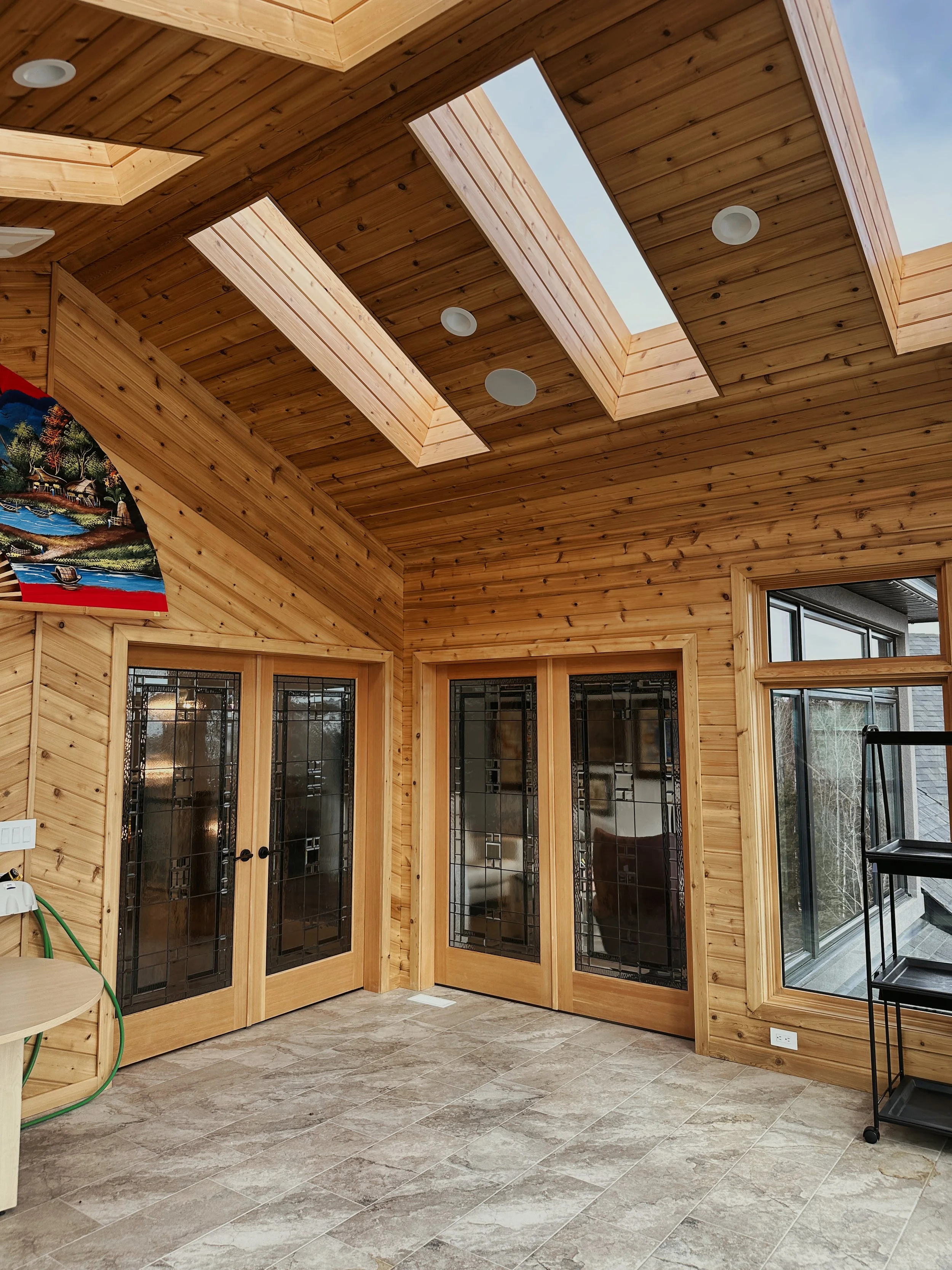 Interior of a cedar room with a vaulted ceiling and skylights, with glass-paneled double doors and a large window, and a black shelving unit on the right.