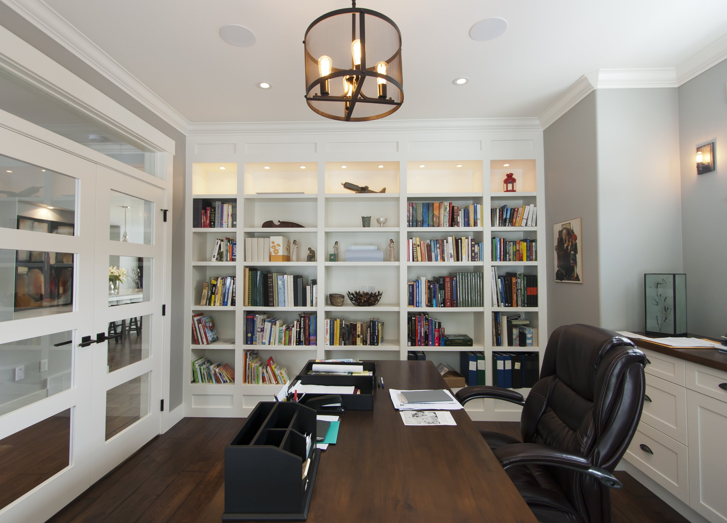 Modern home office with dark wood desk, black leather chair, white bookshelf filled with books and decorative items, gray walls, white trim, and black and gold chandelier.