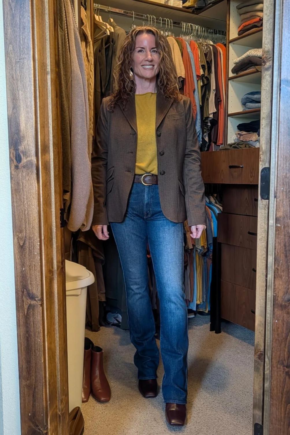 Woman standing in a walk-in closet wearing a brown blazer, yellow shirt, blue jeans, and brown boots, with racks of clothing around her.
