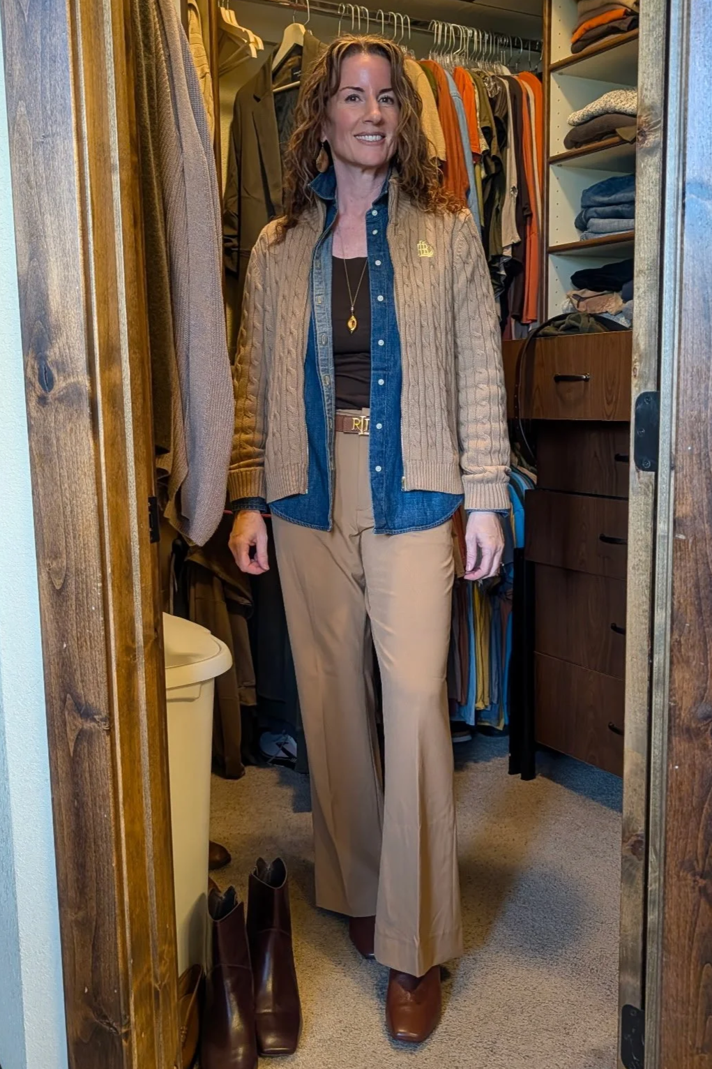 Woman standing in a walk-in closet filled with clothing, wearing beige wide-leg pants, a denim shirt layered with a brown cable-knit cardigan, brown boots, and gold accessories.