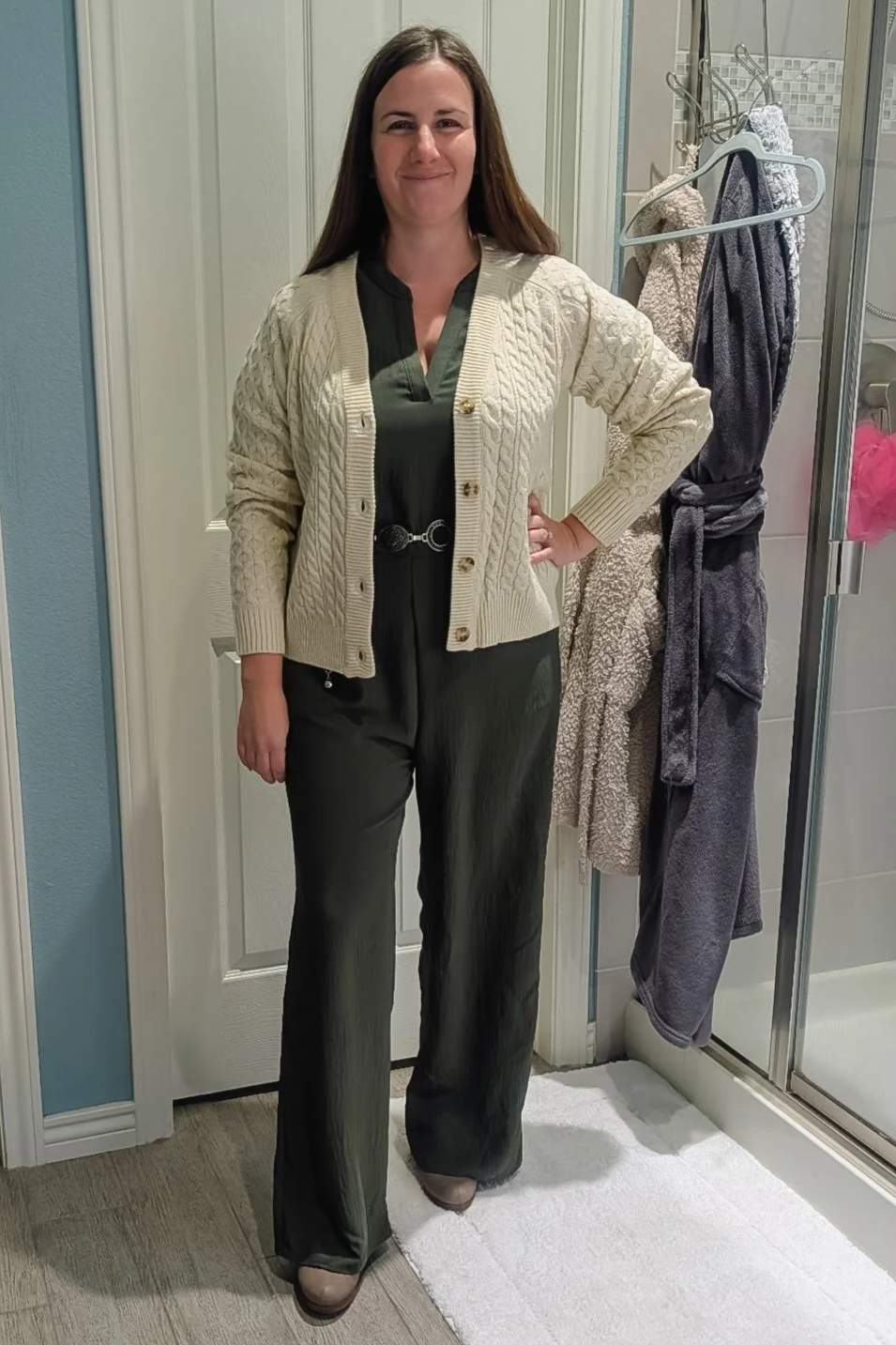 Woman smiling, standing in a bathroom in front of a door, wearing a beige cardigan over a dark top and dark pants.