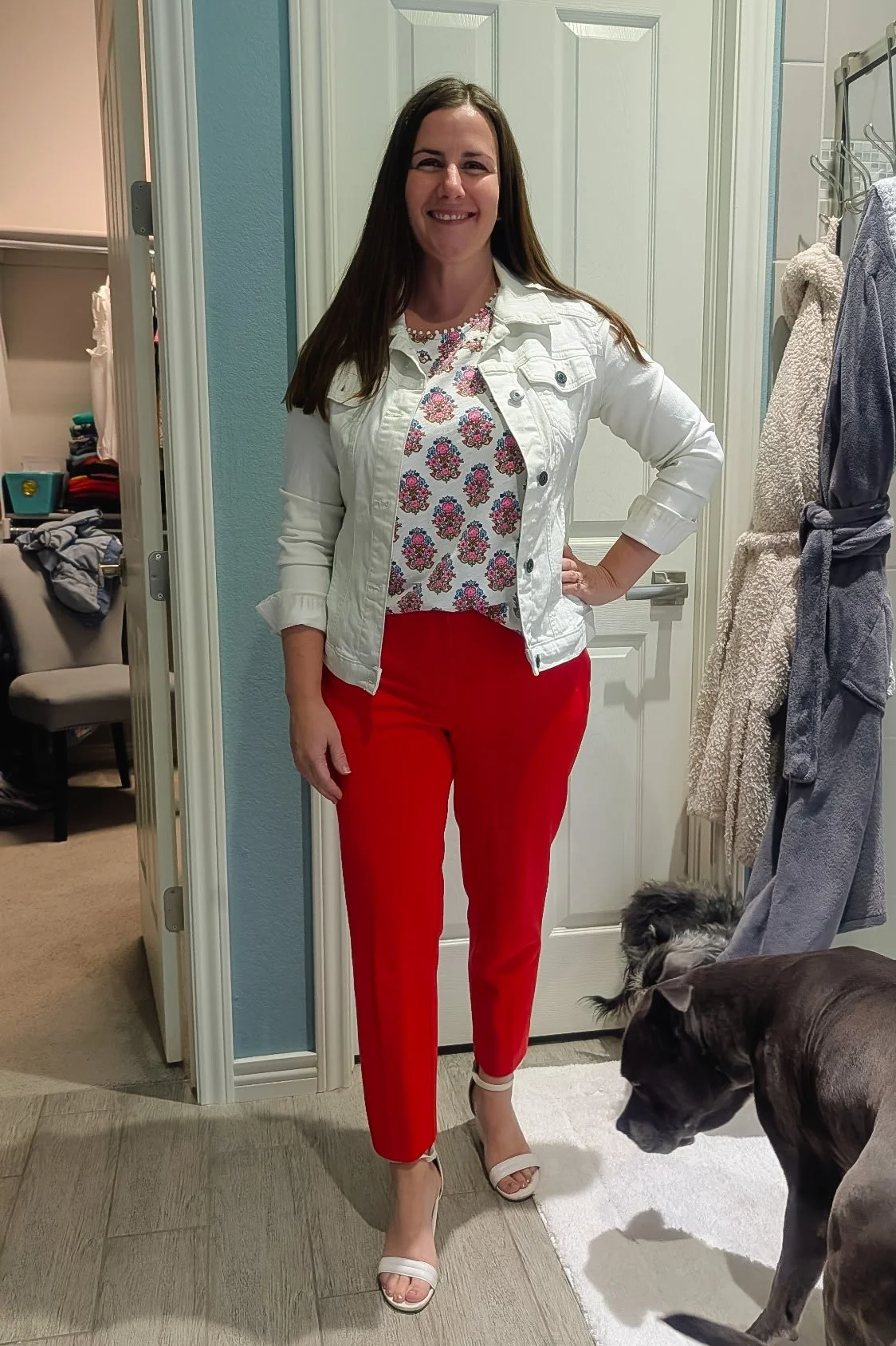 A woman stands in a doorway smiling, wearing a white denim jacket, a floral top, red pants, and white sandals. Two dogs are nearby, one black and one gray, with a room and closet in the background.