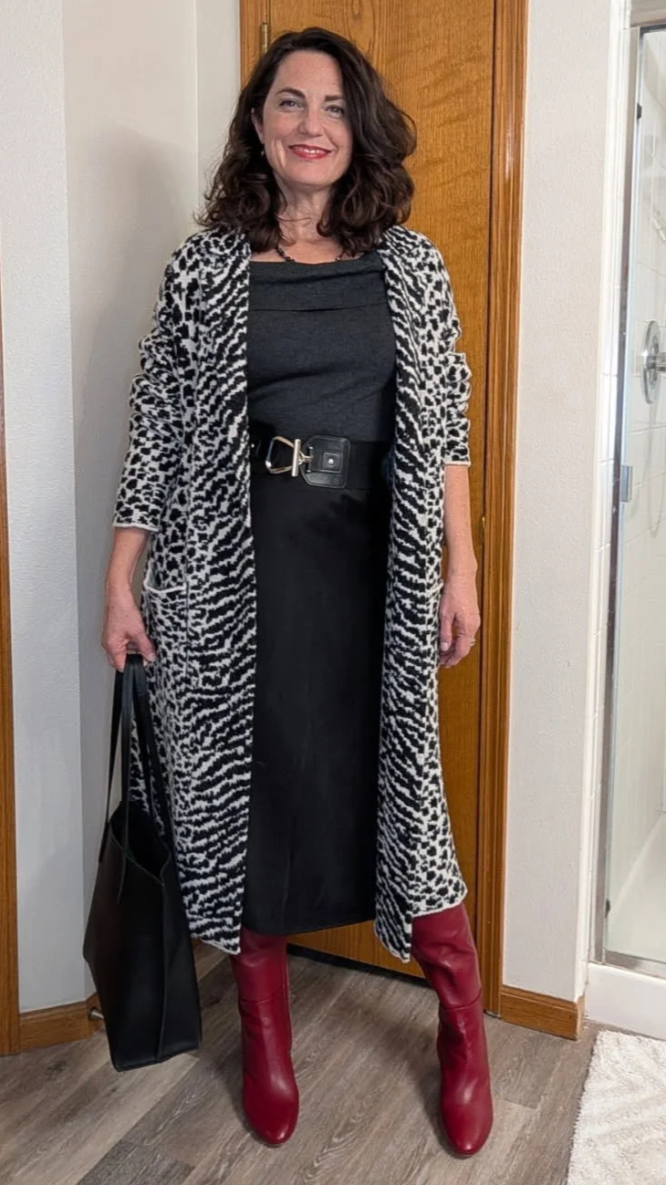 A woman with shoulder-length dark hair, wearing a black top, a leopard print cardigan, a black skirt with a wide belt, and red knee-high boots, standing indoors near a wooden door and a glass shower door.