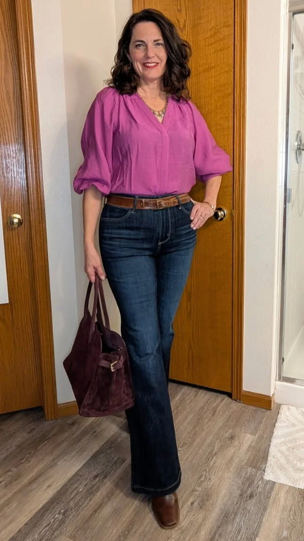 A woman with dark, curly hair wearing a pink blouse, dark blue jeans with a brown belt, and brown shoes, holding a dark purple bag, standing in a room with wooden doors and a wooden floor.