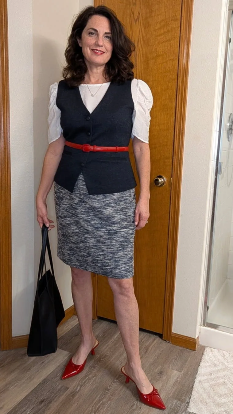 A woman with shoulder-length curly brown hair, wearing a white puff-sleeve blouse with a black vest and matching skirt, red belt, red high heels, holding a black bag, standing in a room with wooden door and beige walls.