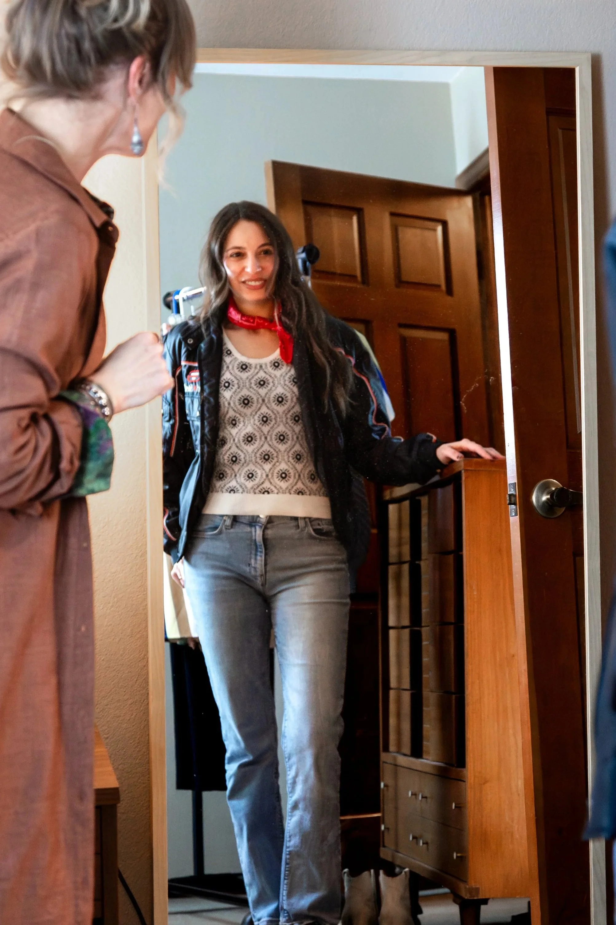 A young woman with long dark hair, wearing a patterned top, jeans, and a dark jacket, stands in front of a mirror smiling at her reflection.