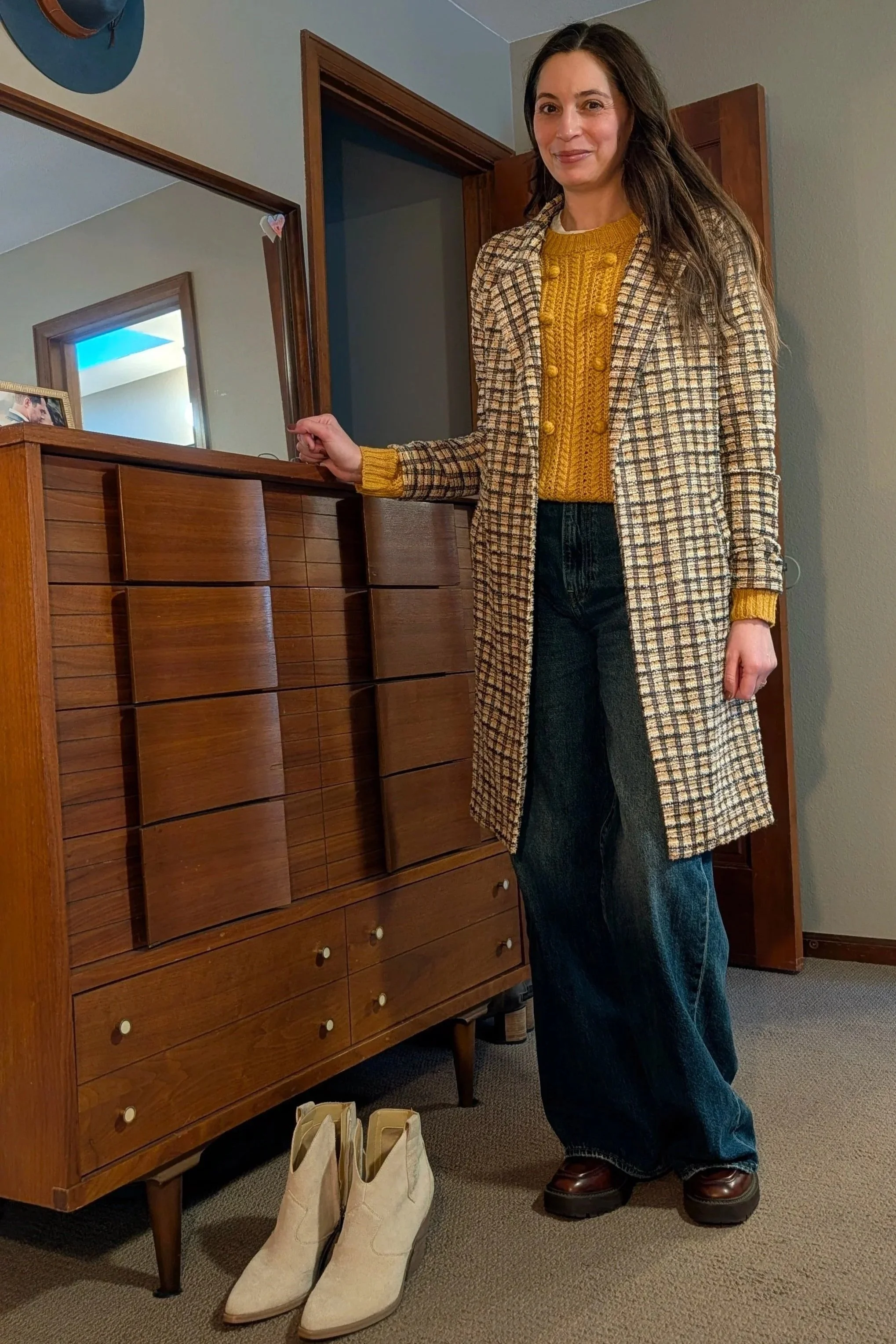 A woman standing next to a wooden dresser with a mirror, wearing a yellow sweater, plaid coat, wide-leg jeans, and brown boots, in a room with beige walls and a mirror reflection.