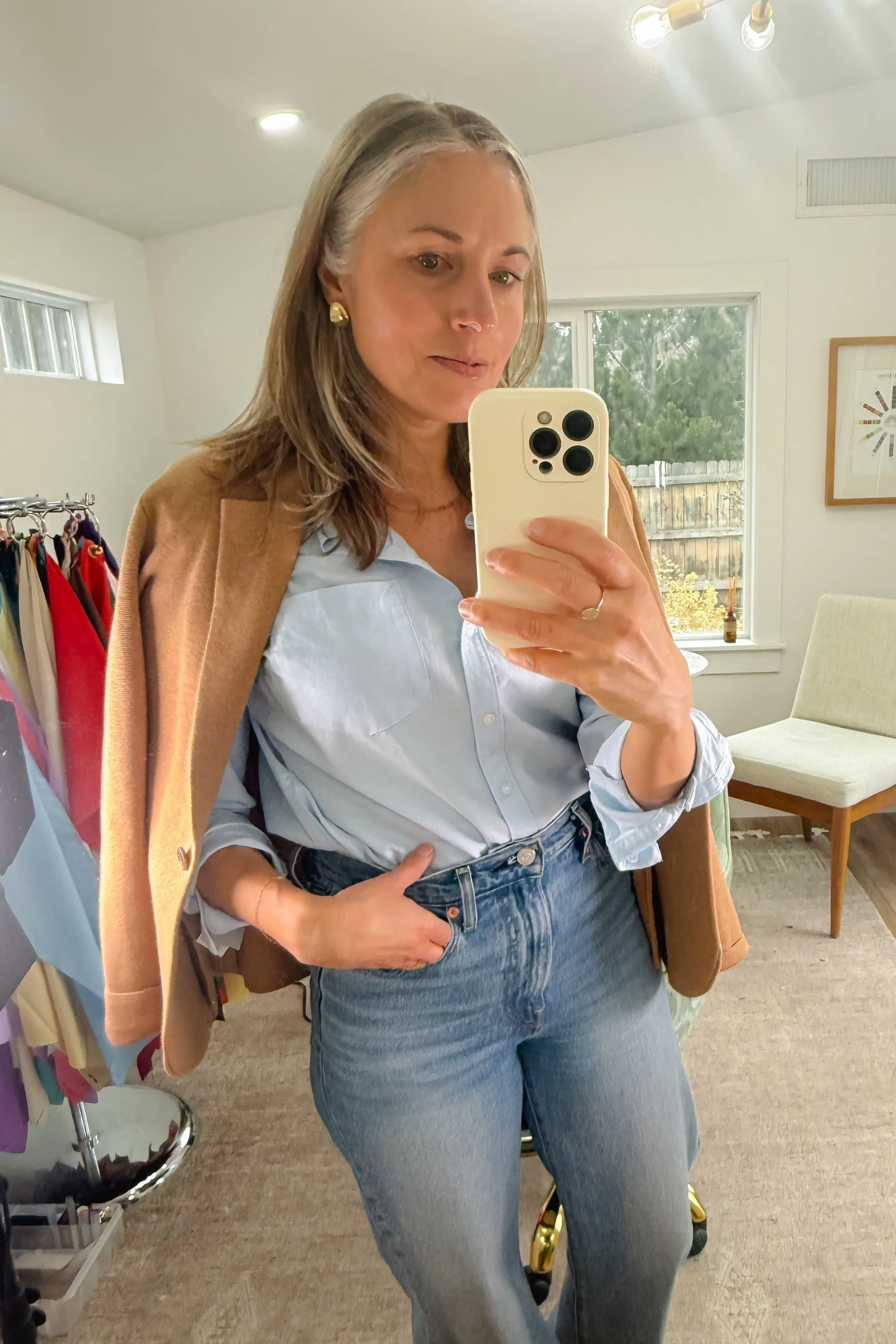 Woman taking a mirror selfie with her phone, wearing a light blue button-up shirt and high-waisted jeans, with a brown blazer draped over her shoulders, in a room with a window, beige chair, and clothing rack.