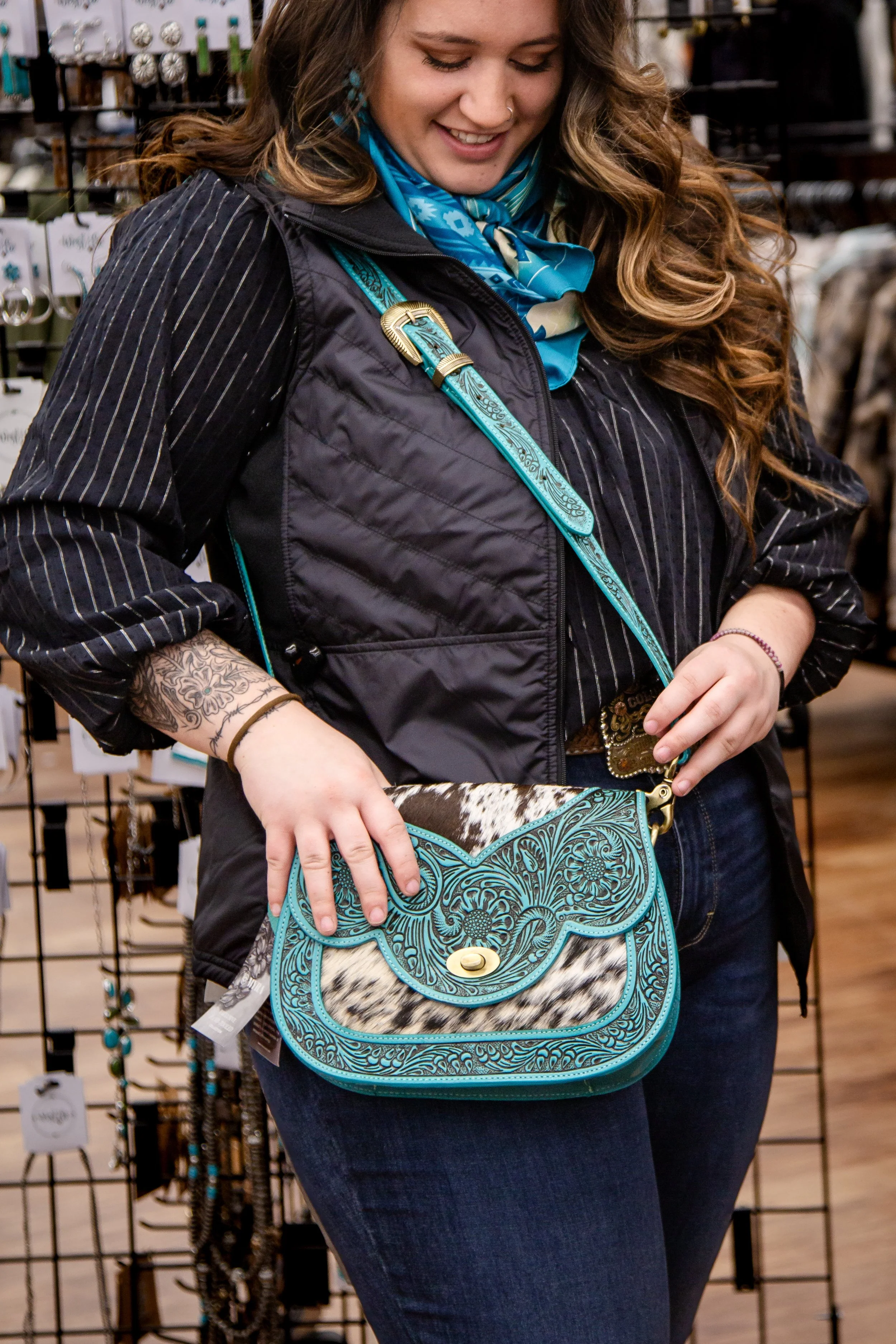 A woman with long, curly hair wearing a black pinstripe blazer and a blue bandana is shopping for accessories in a store. She has a tattoo on her left wrist and is holding a turquoise and cowhide patterned handbag. She is smiling and looking down, wi