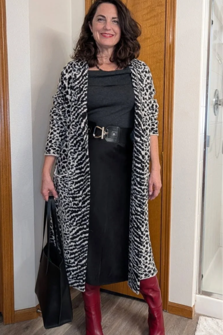 A young woman with long, dark hair smiling outdoors against a bright blue sky, wearing a black floral blouse and dark jeans.
