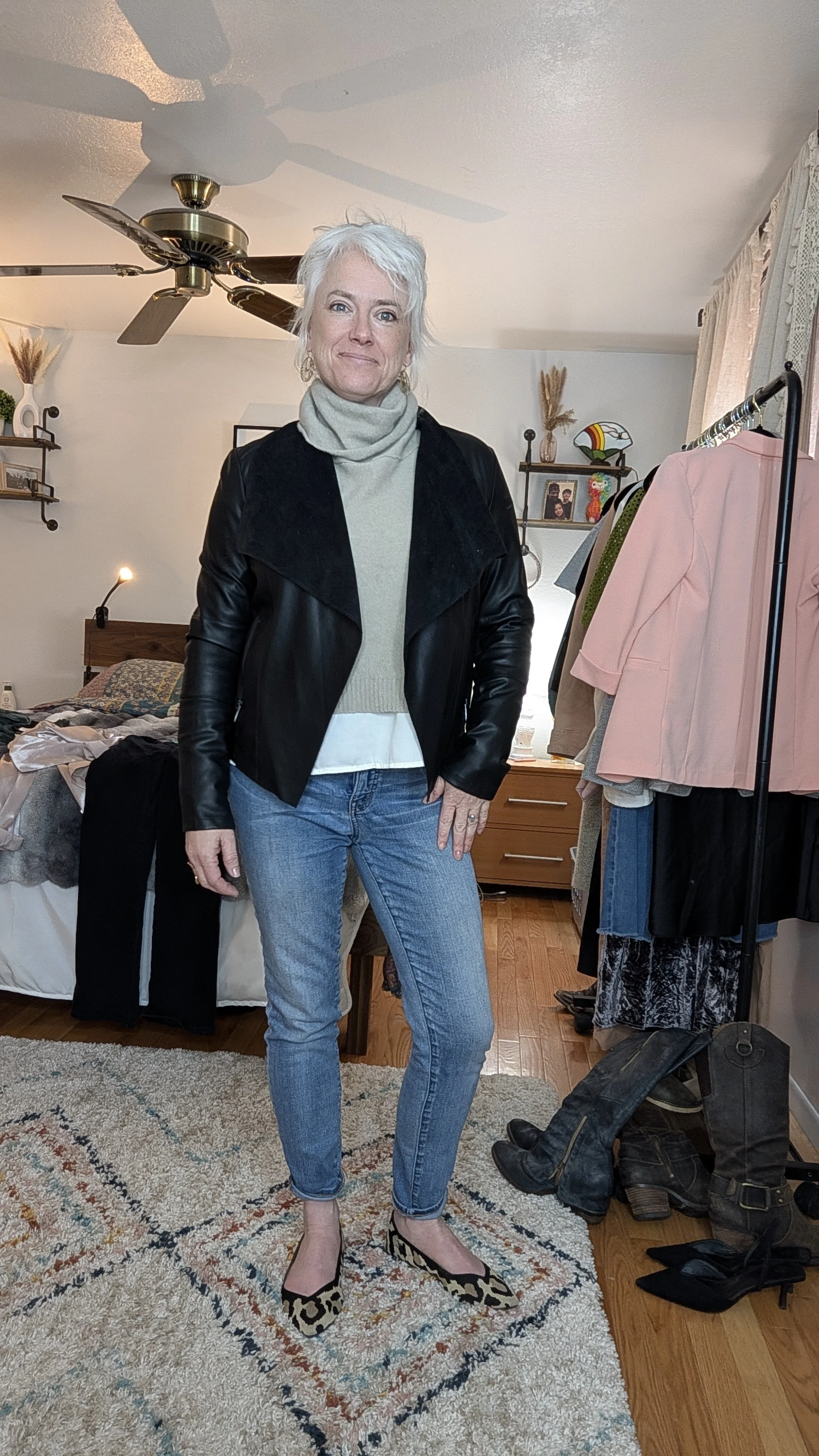 A woman with short white hair standing in a bedroom, wearing a gray turtleneck, black leather jacket, blue jeans, and leopard print shoes. The room has a bed, clothing rack, and various decorations.