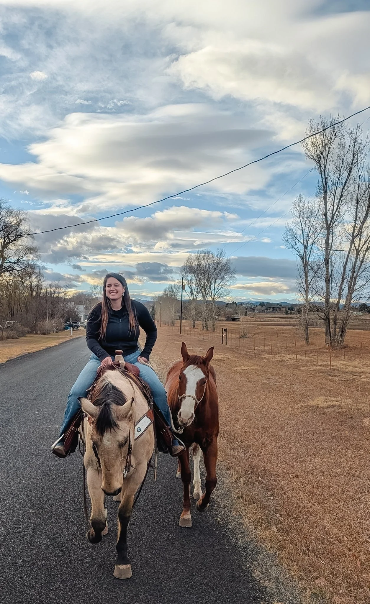 A woman riding a beige horse on a paved country road with a brown and white pinto horse walking beside her, under a partly cloudy sky with leafless trees and open fields in the background.