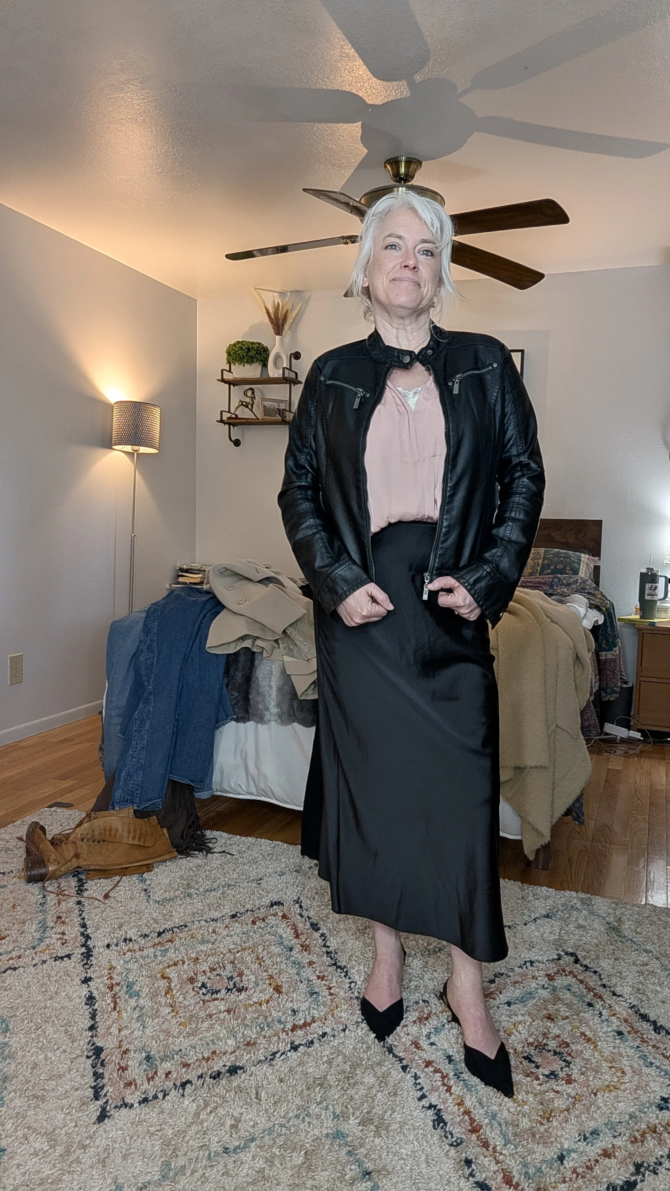 A woman with white hair and light skin standing in a bedroom. She is wearing a black leather jacket, a light pink blouse, a black skirt, and black high heels. The room has a bed with clothes on it, a nightstand, and a ceiling fan.