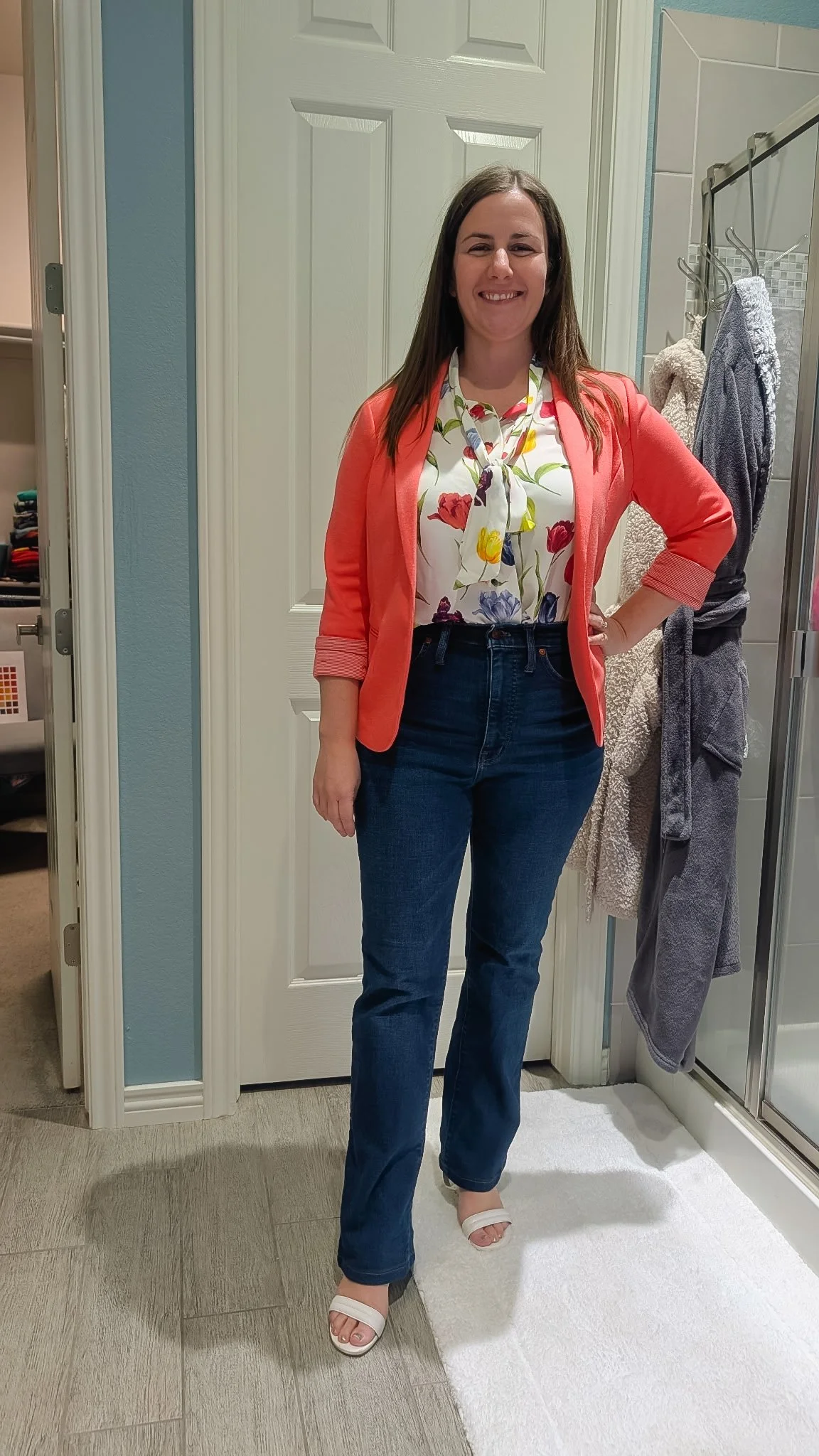 A woman standing in a bathroom, wearing a floral blouse, coral blazer, dark jeans, and white sandals, smiling at the camera.