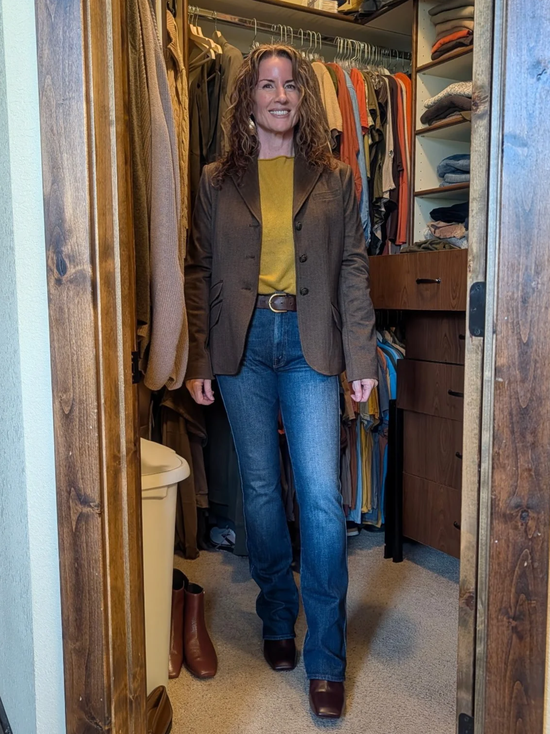 A woman standing outdoors against a dark wall with greenery, wearing a beige coat over a green dress and high heels.