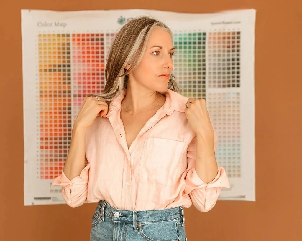 A woman with shoulder-length blonde hair wearing a light pink blouse and jeans standing in front of a color map poster on a brown wall.