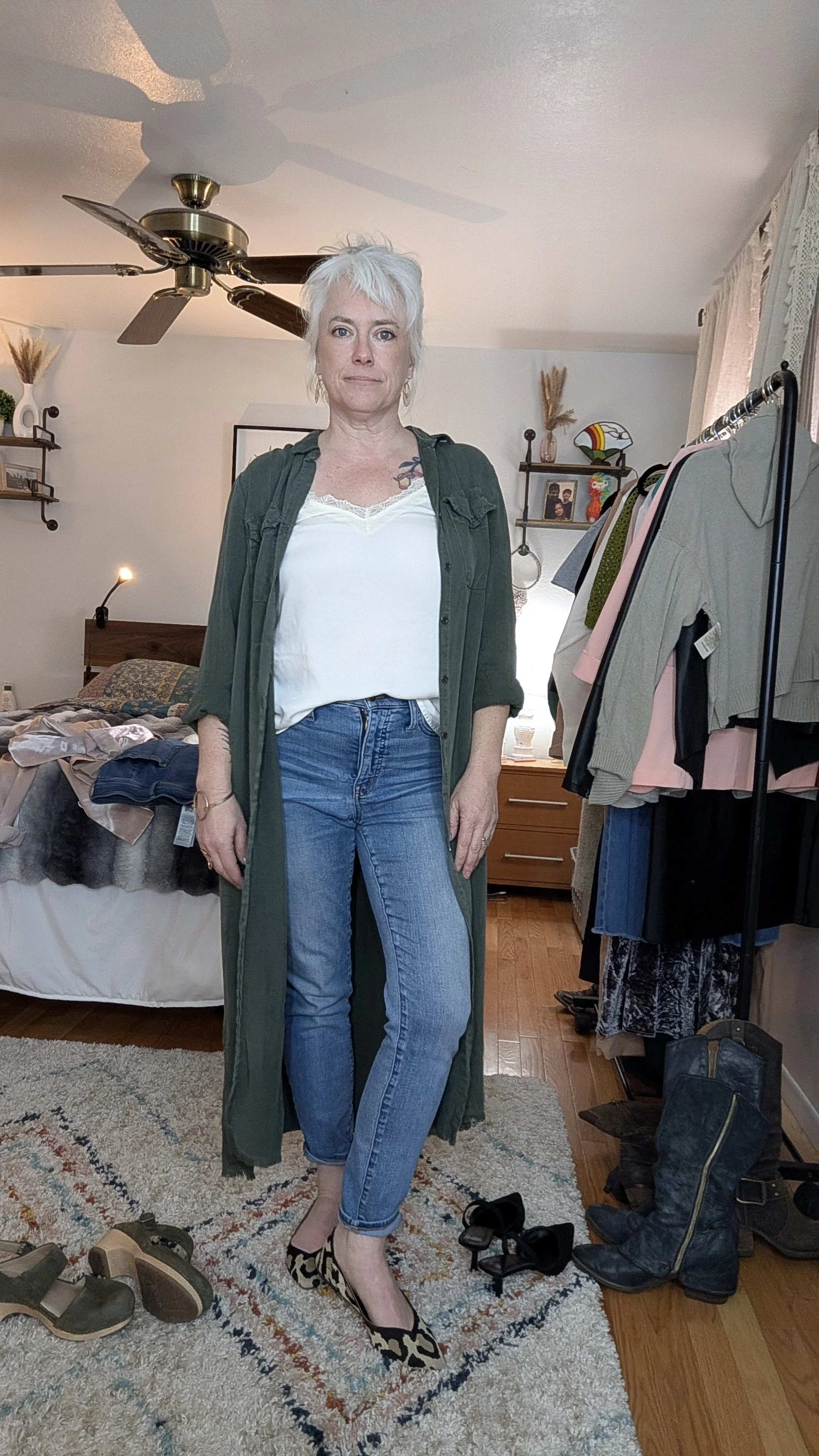 A woman with short white hair standing in a bedroom, dressed in a white top, blue jeans, leopard print heels, and a long green coat. The room has a bed, a ceiling fan, a clothing rack with various clothes, and a pair of black cowboy boots on the floo