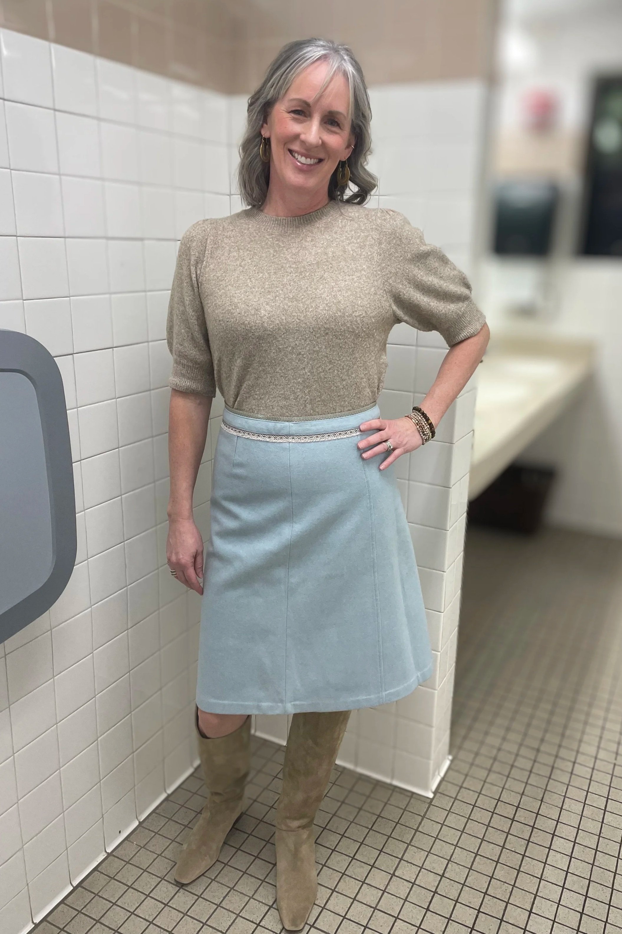 A woman with gray hair smiling, wearing a beige sweater, light blue skirt, beige knee-high boots, and jewelry, standing in a restroom with white tiled walls and a beige counter.