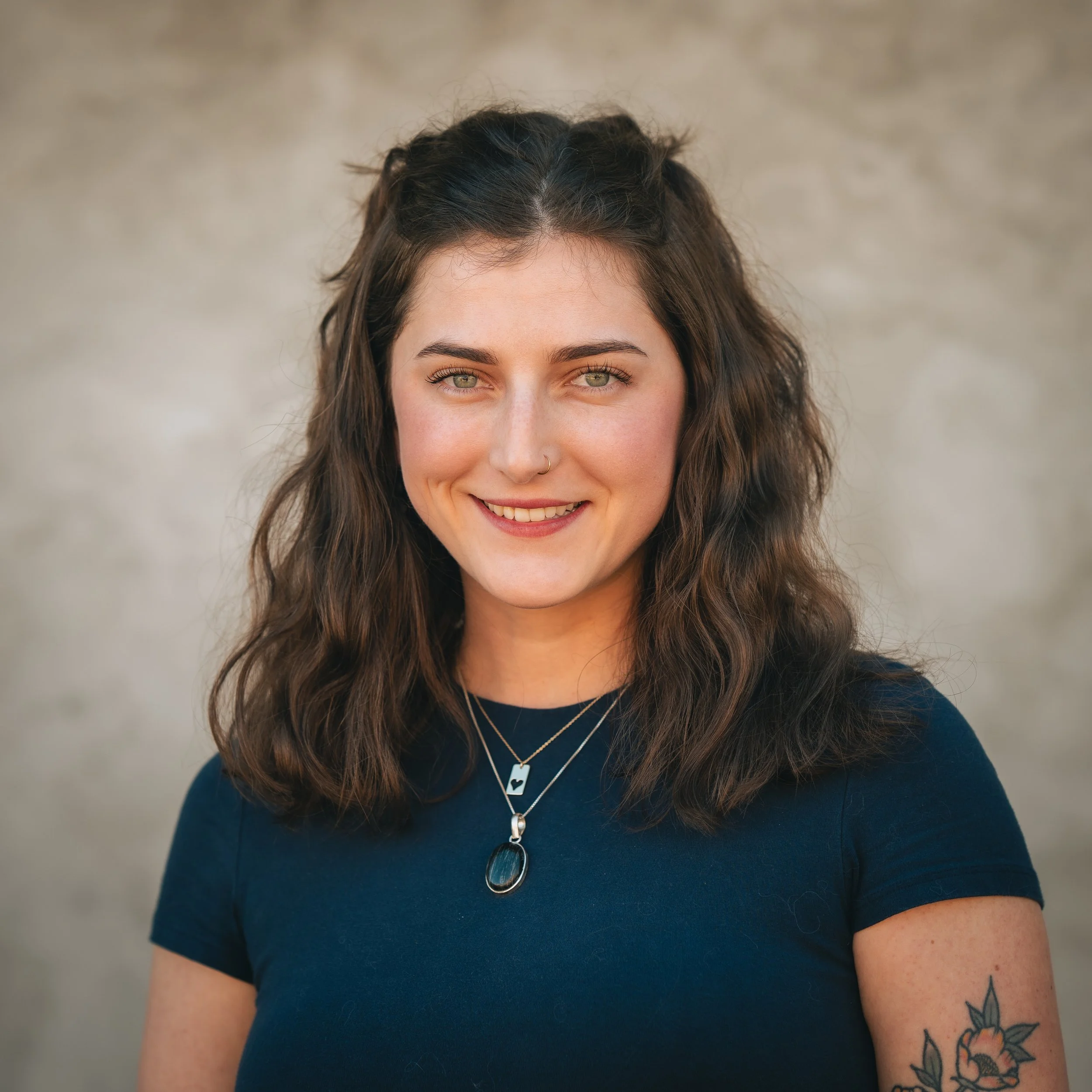 Portrait of a young woman with wavy brown hair, wearing a dark blue shirt, layered necklaces, and a floral tattoo on her left arm, smiling against a neutral background.
