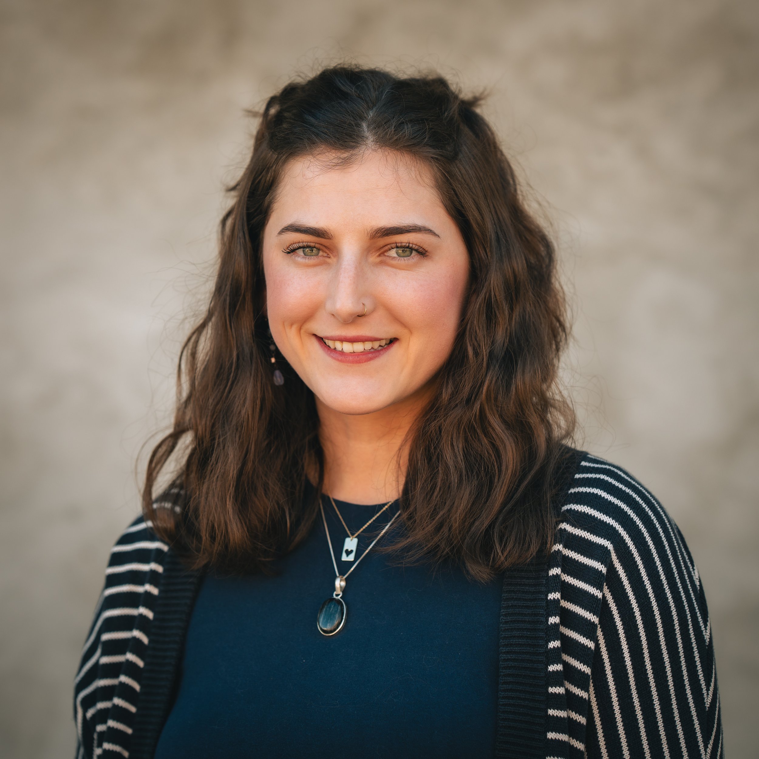A young woman with wavy brown hair and green eyes, smiling, wearing a navy blue top, striped cardigan, layered necklaces, and earrings, standing outdoors with a blurred neutral background.