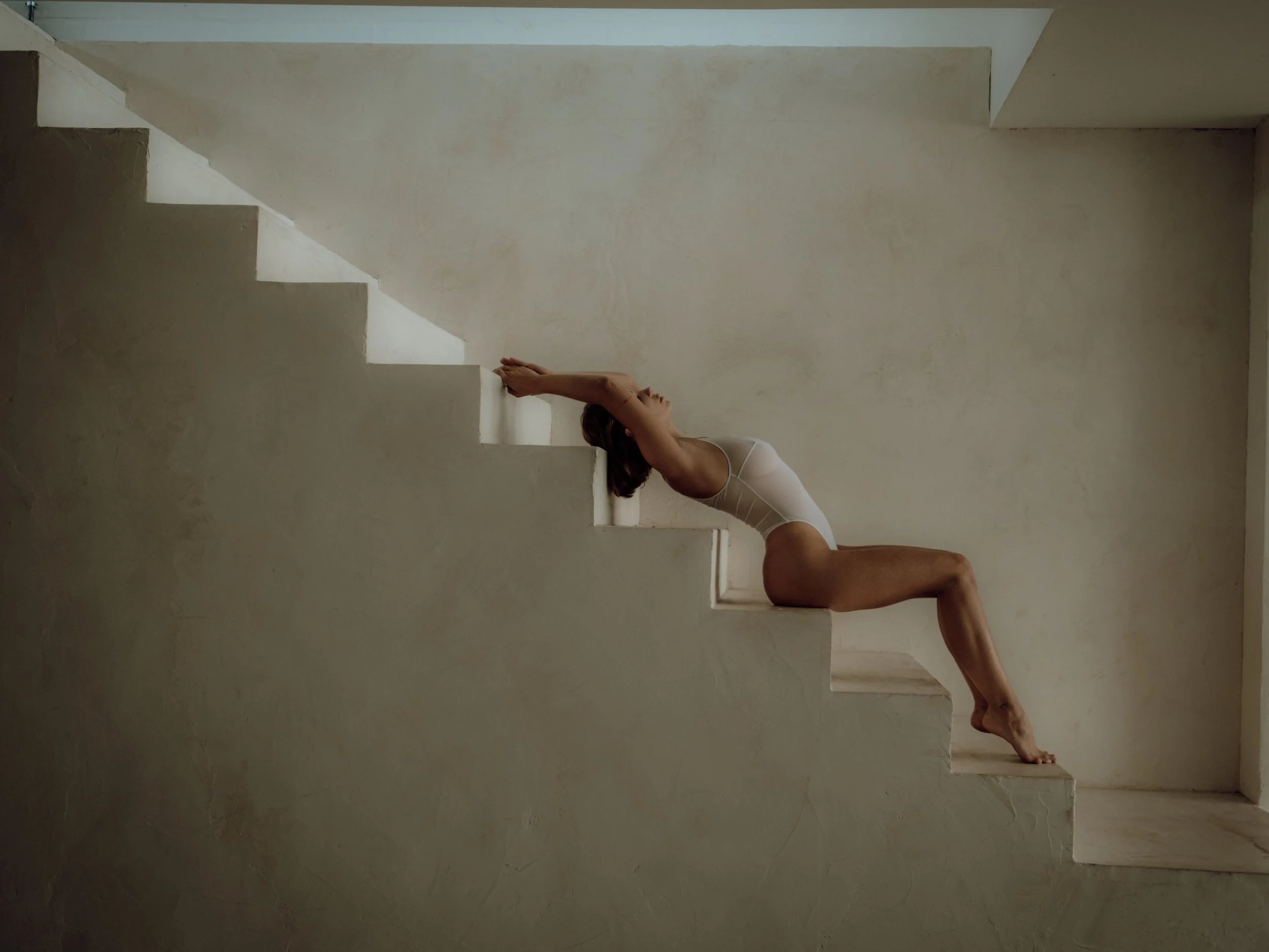 A woman in a white bodysuit performing a yoga pose on a staircase with a plain beige wall in the background.