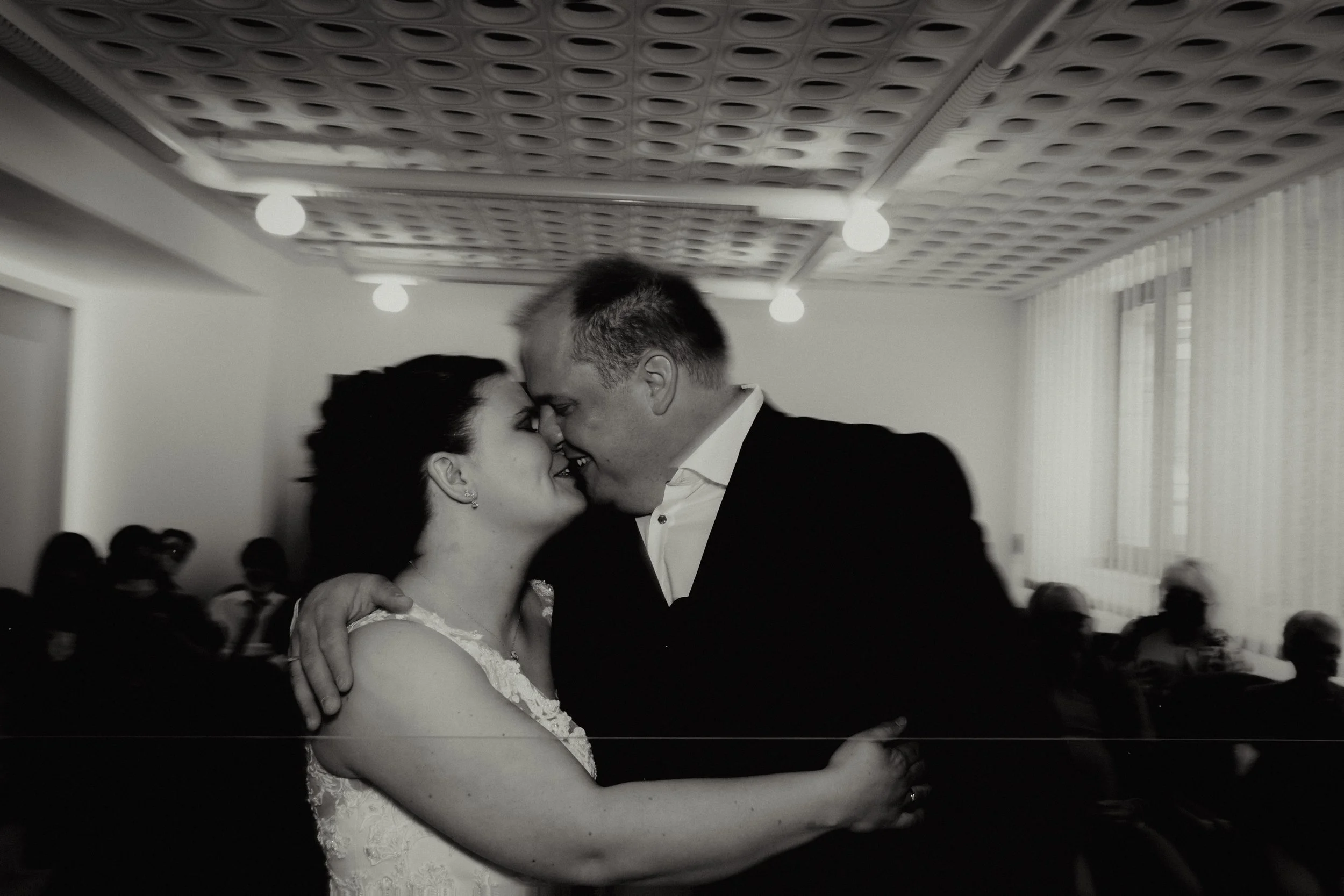 A black and white photo of a couple dancing closely, with their foreheads touching and smiling, in a room with bright lighting and other people seated in the background.