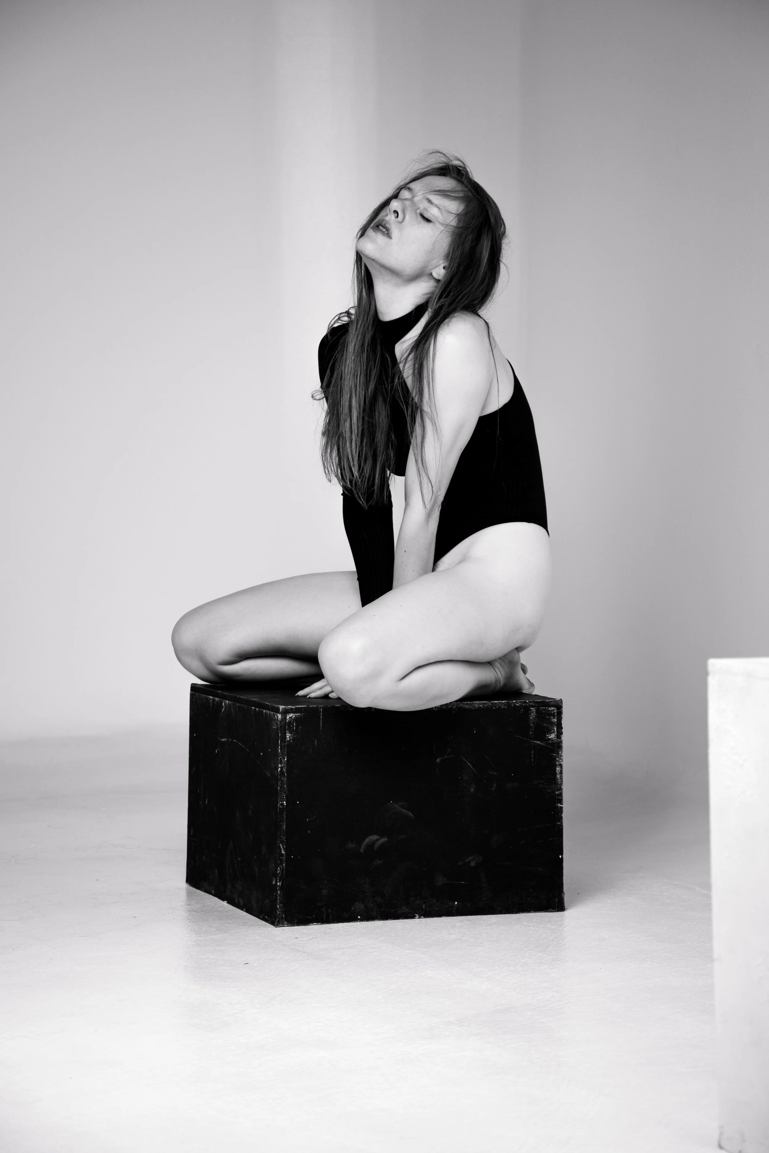 A woman kneeling on a black cube in a minimalistic space, wearing black clothing and looking upward with her hair falling over her face, in black and white photography.