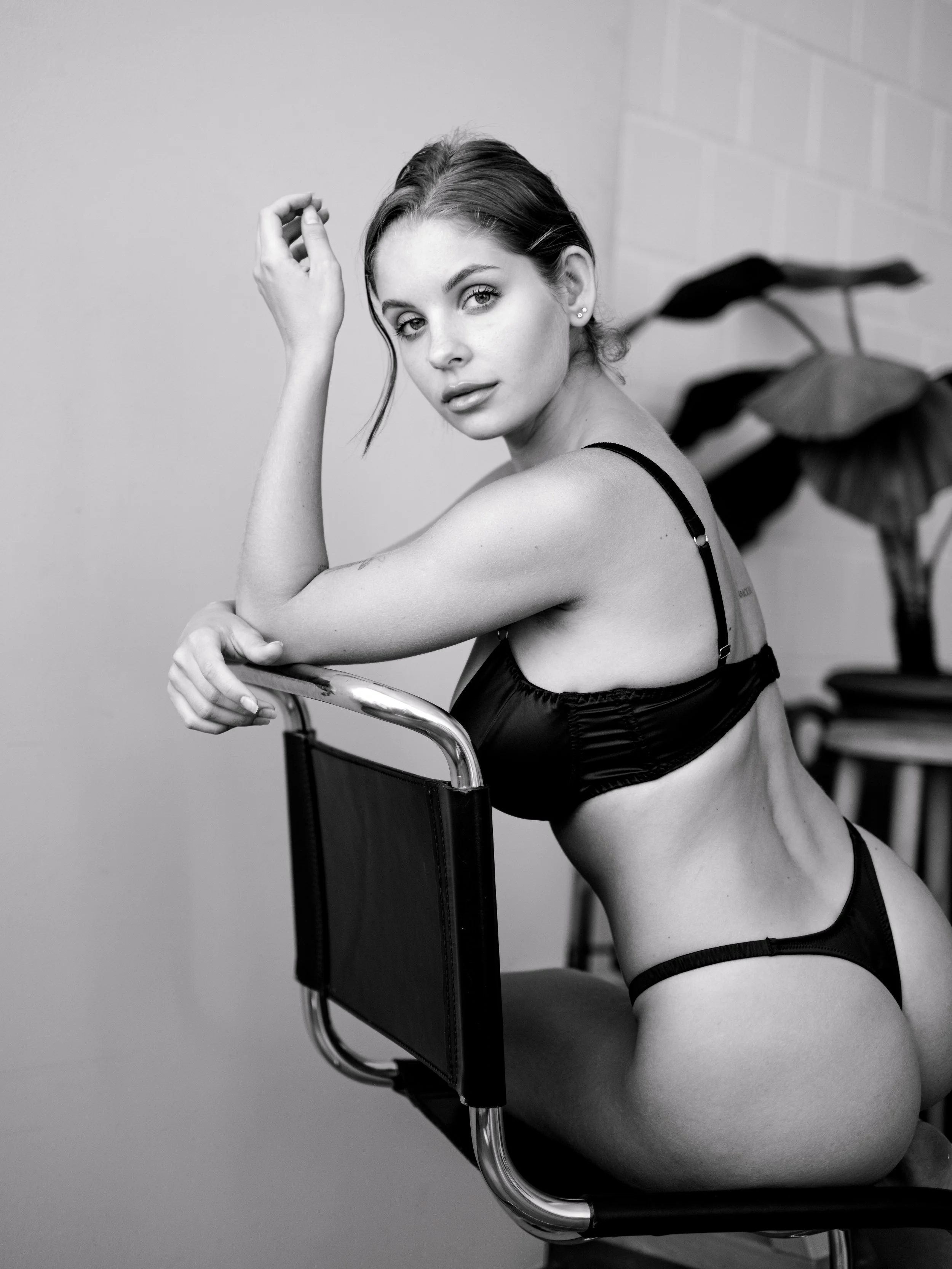Black and white photo of a woman sitting on a chair, leaning forward with her arms resting on a chair in front of her, looking at the camera.