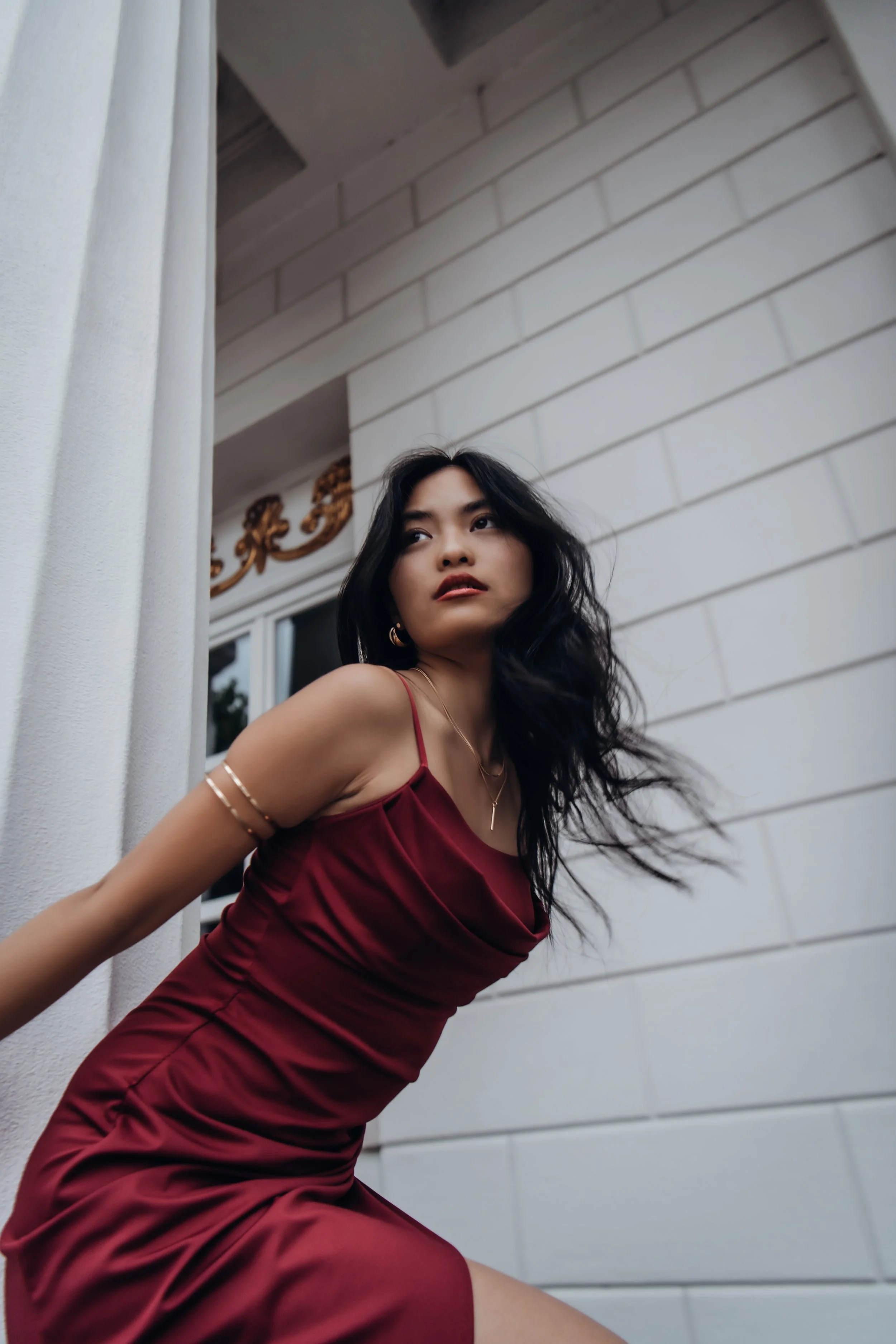 A woman with long wavy black hair wearing a satin red dress with thin straps, leaning against a white wall outdoors with decorative gold accents, looking to the side.