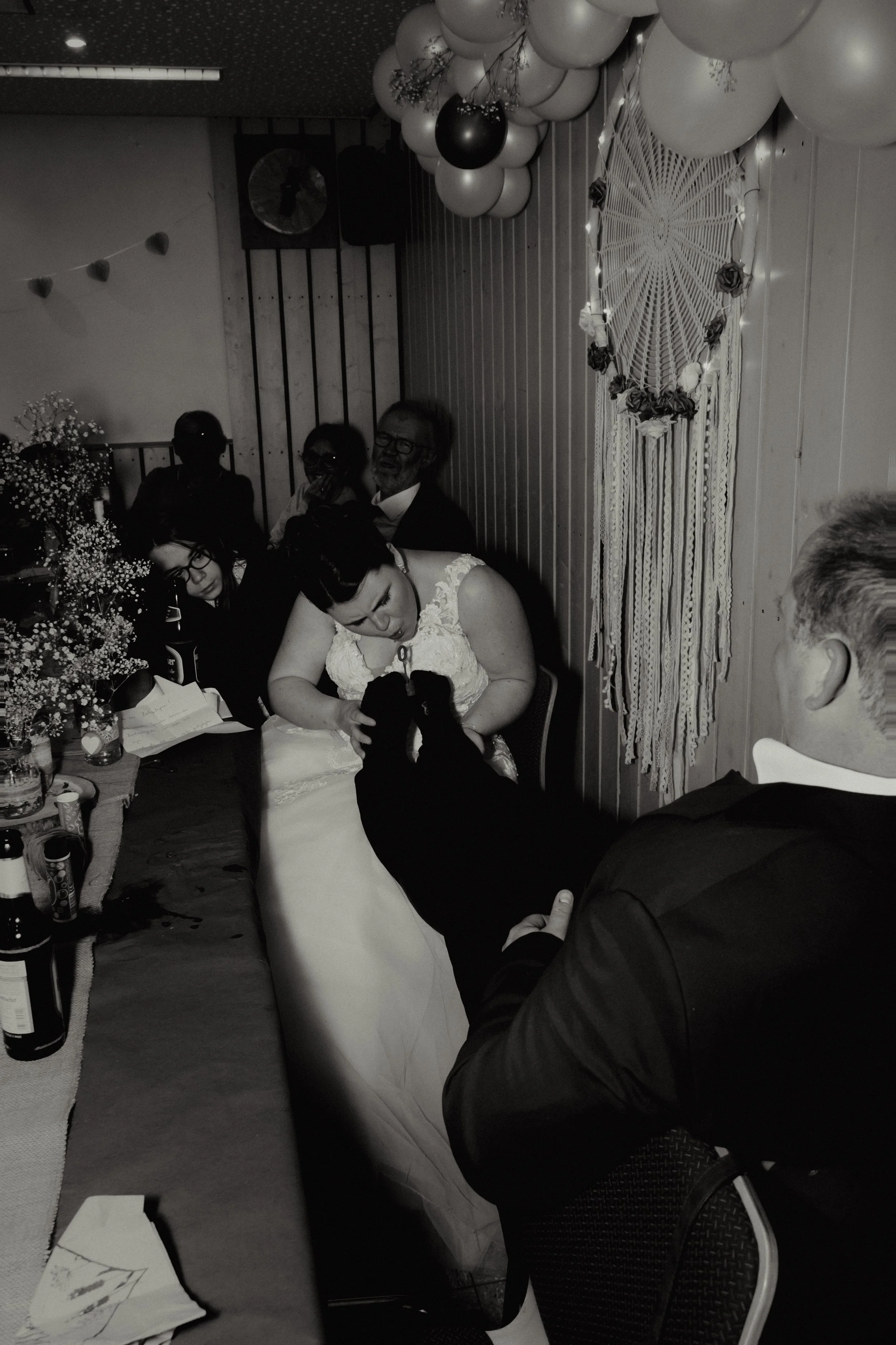 A bride in a wedding dress is kissing a black dog on her lap at a wedding reception, surrounded by guests seated at a decorated table with balloons and flowers.