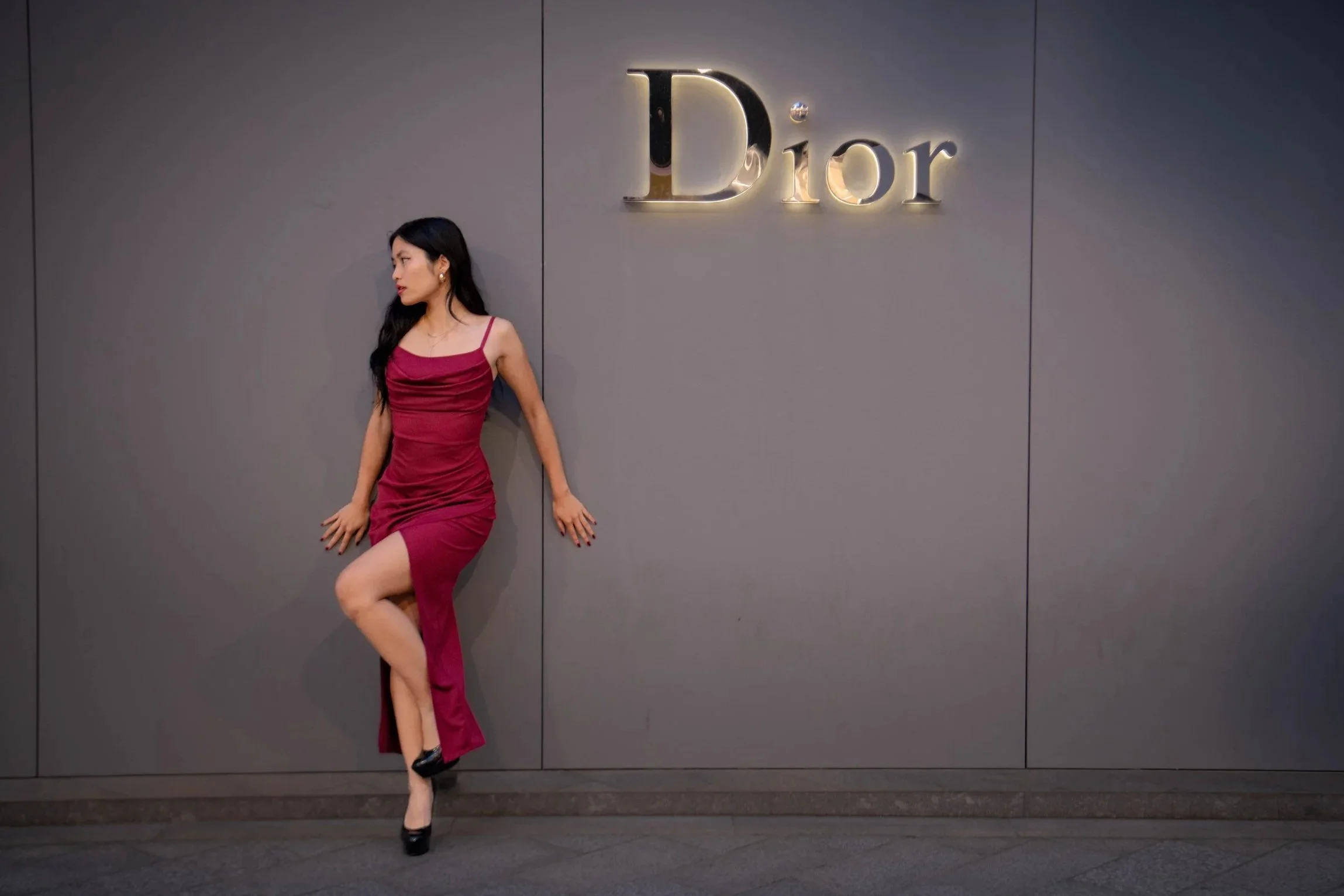 A woman in a red, sleeveless dress and high heels standing against a gray wall with a 'Dior' sign, posing with one leg raised and leaning against the wall.