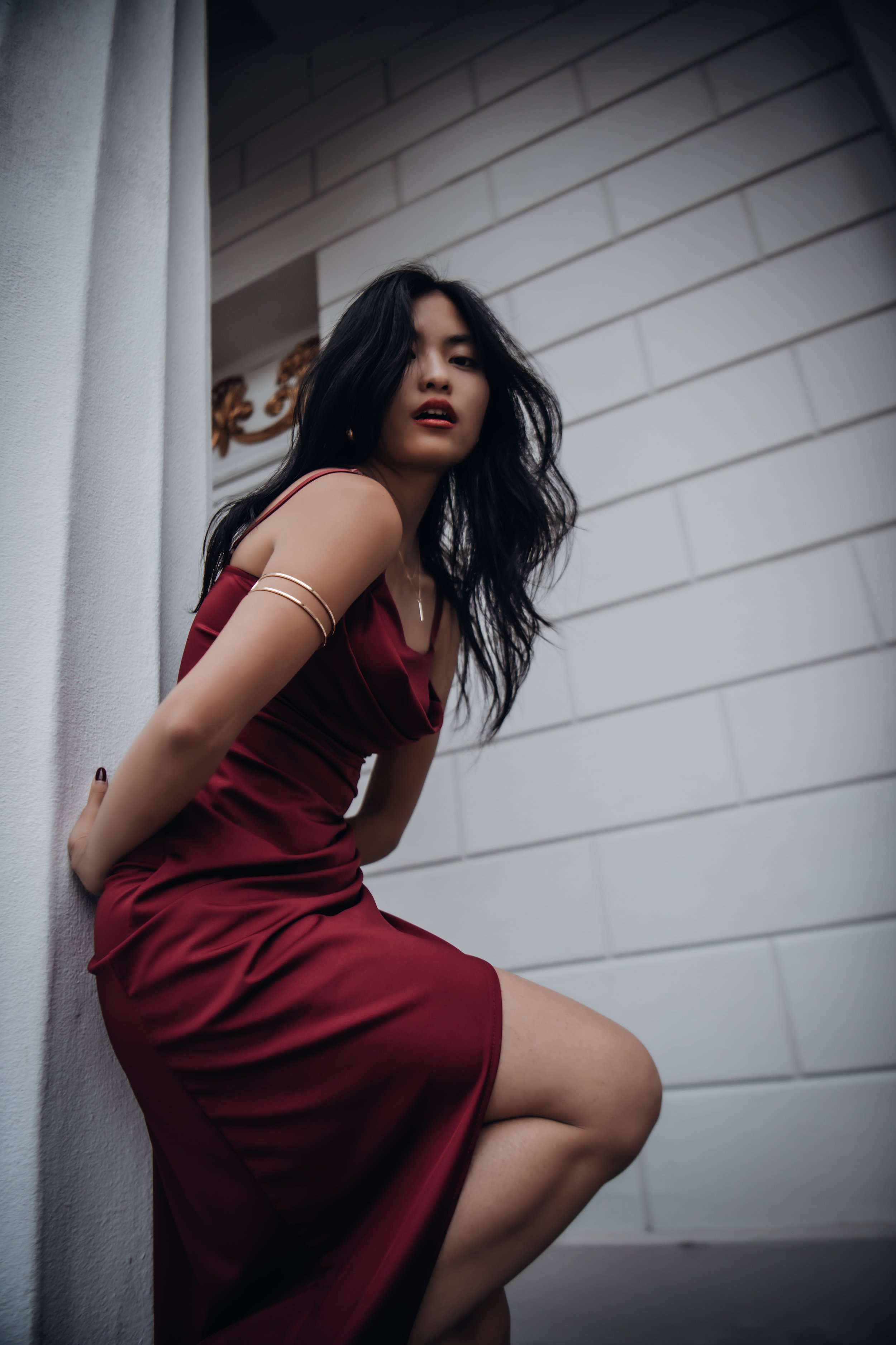 A woman in a red dress crouches against a wall with a white brick pattern, looking at the camera with a serious expression.