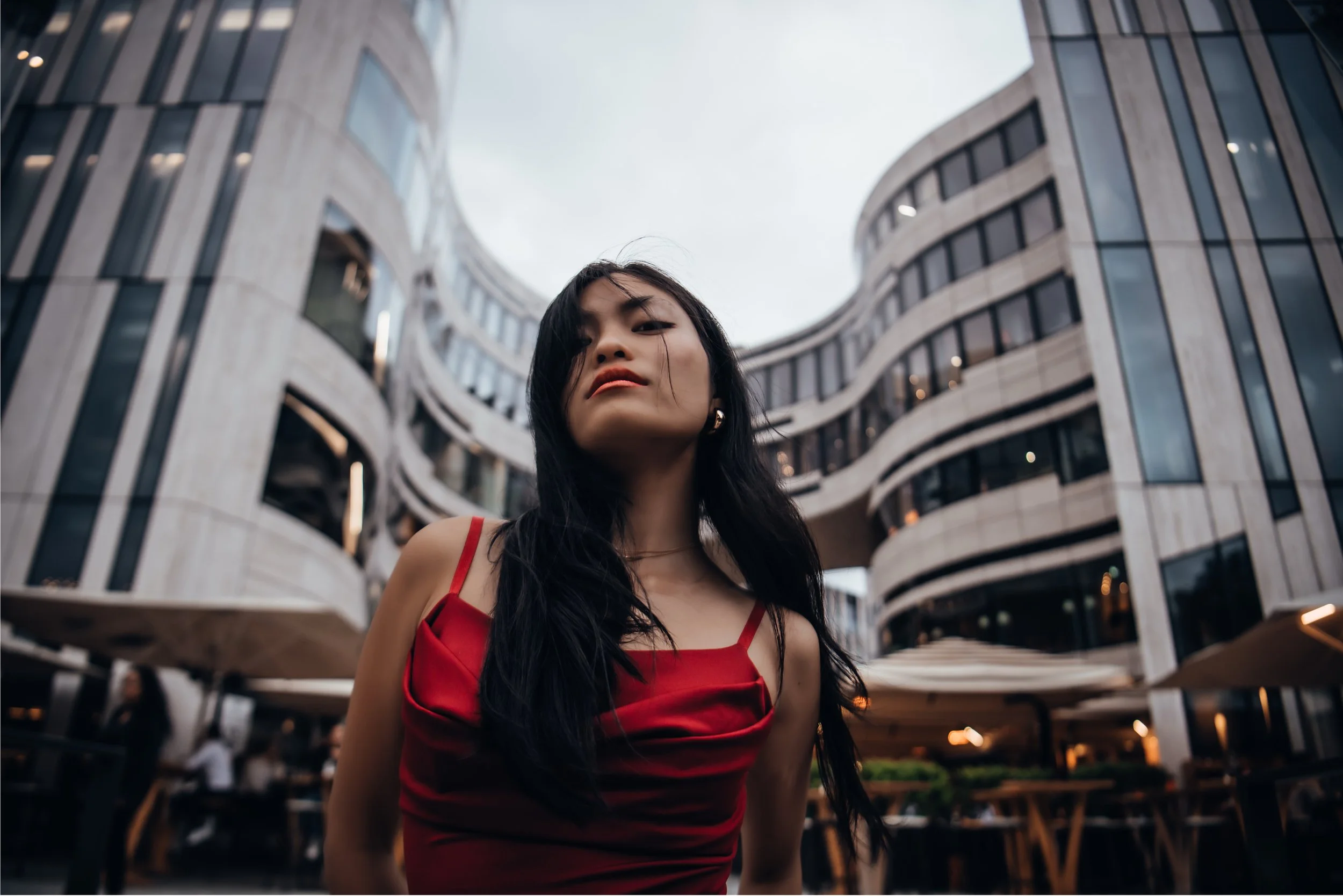 A woman with long dark hair wearing a red dress standing outside in front of a modern, curved multi-story building with glass windows.