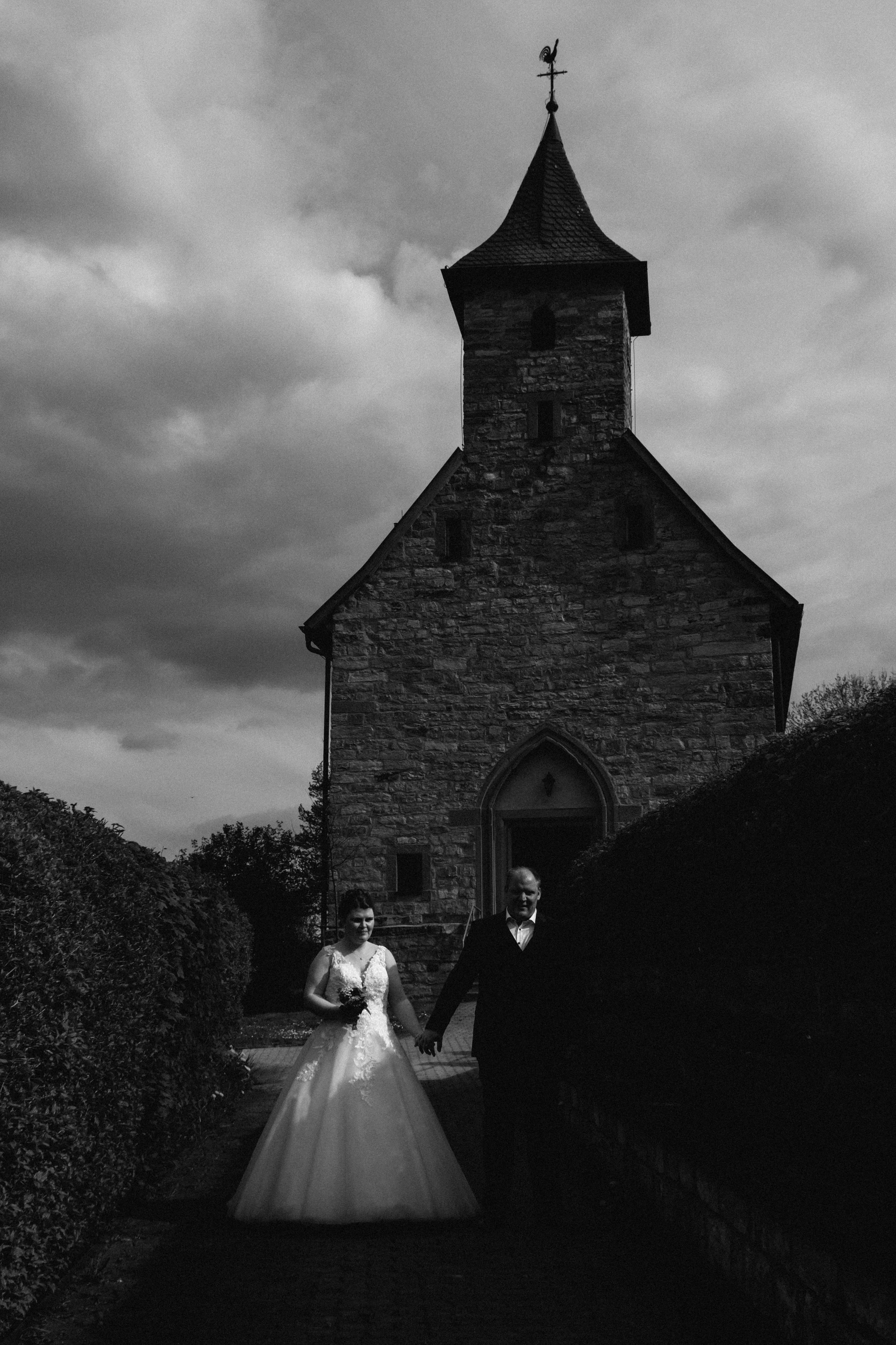 A bride and groom holding hands walking in front of a small stone church with a pointed steeple, cloudy sky in the background.
