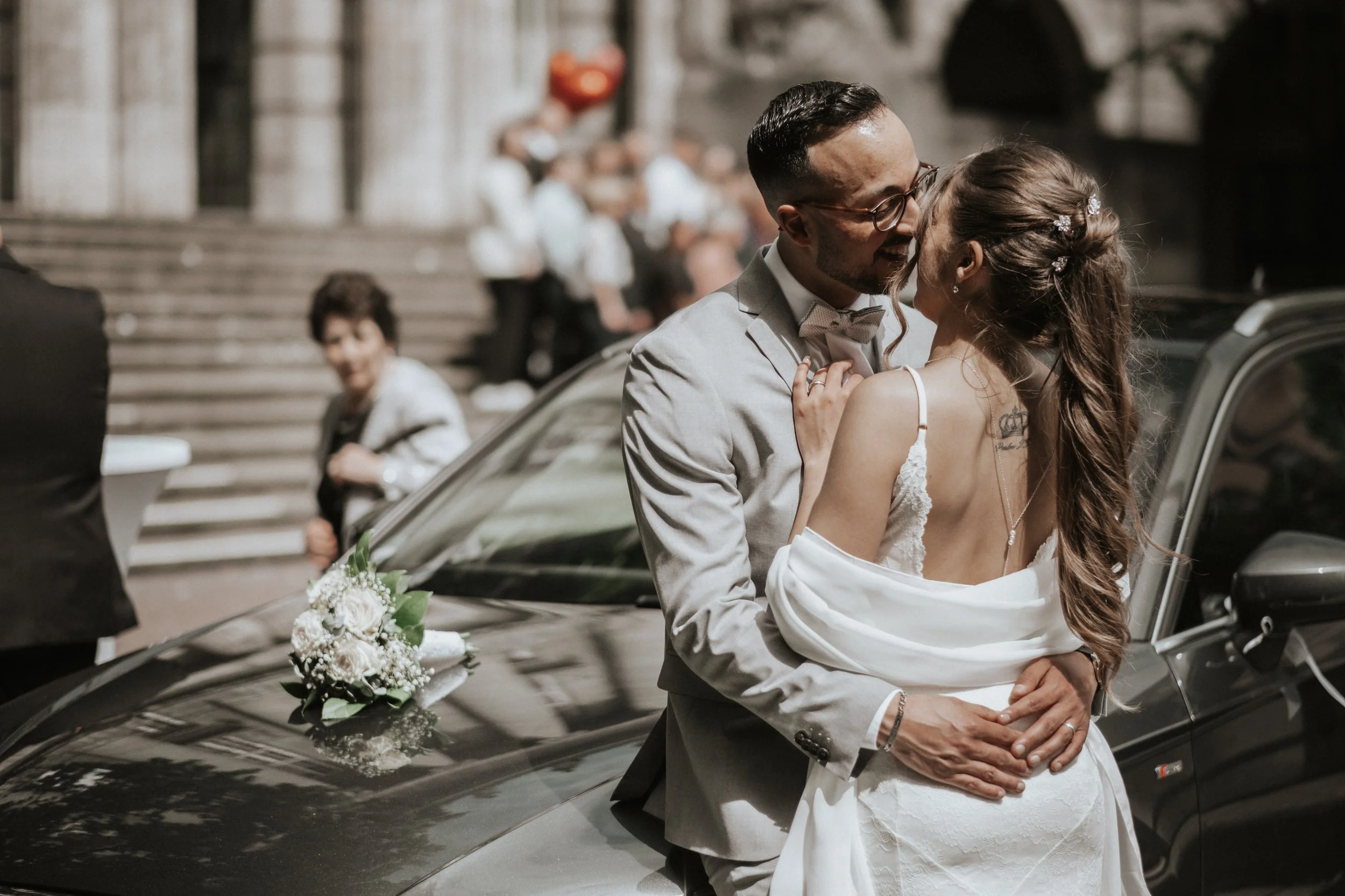 A newlywed couple embracing each other in front of a black luxury car decorated with white flowers, on a city street with steps and onlookers in the background.