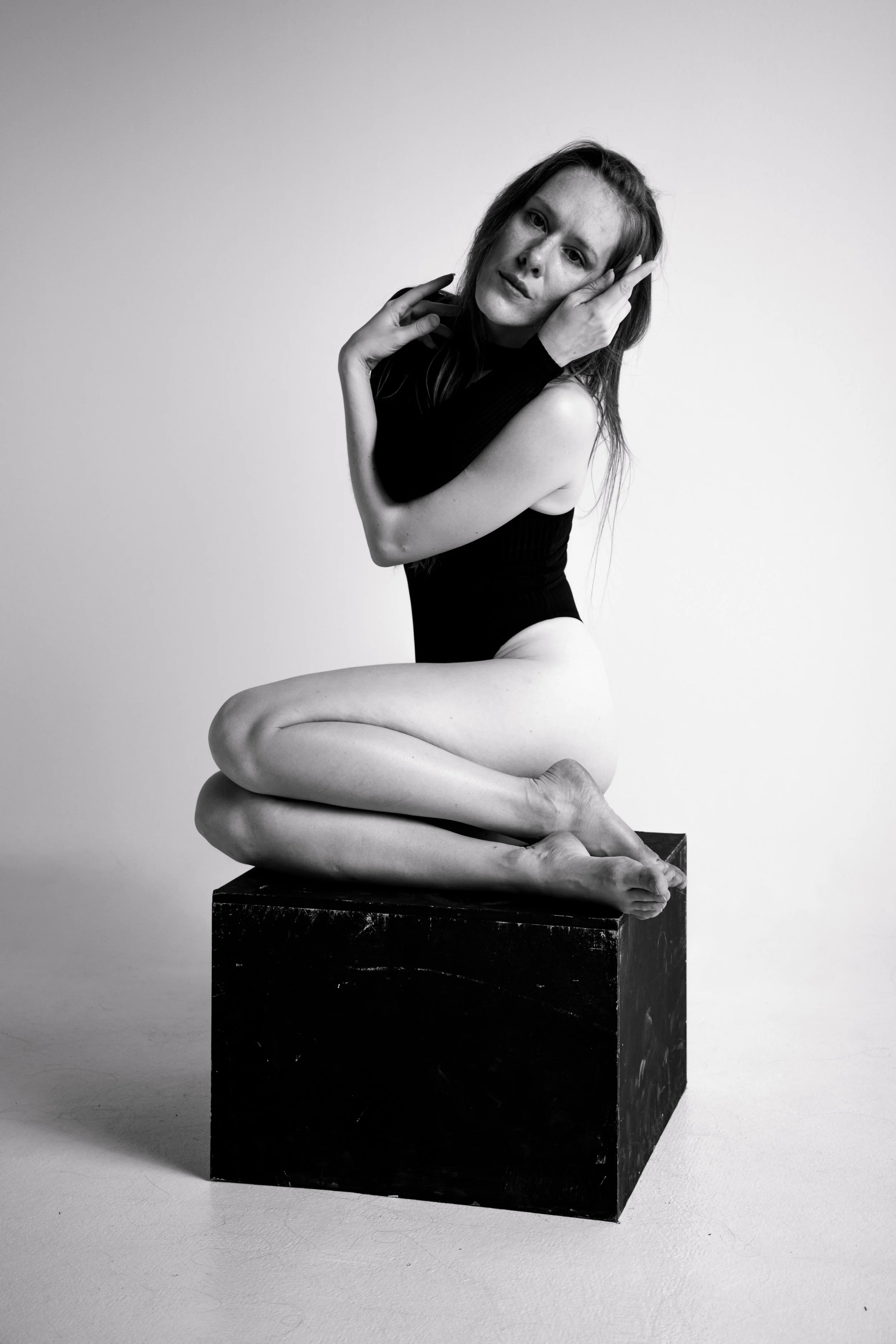 Black and white photo of a woman with long hair, wearing a sleeveless top and arm warmers, sitting on a cube, with her legs crossed and head resting on her hand, looking at the camera.