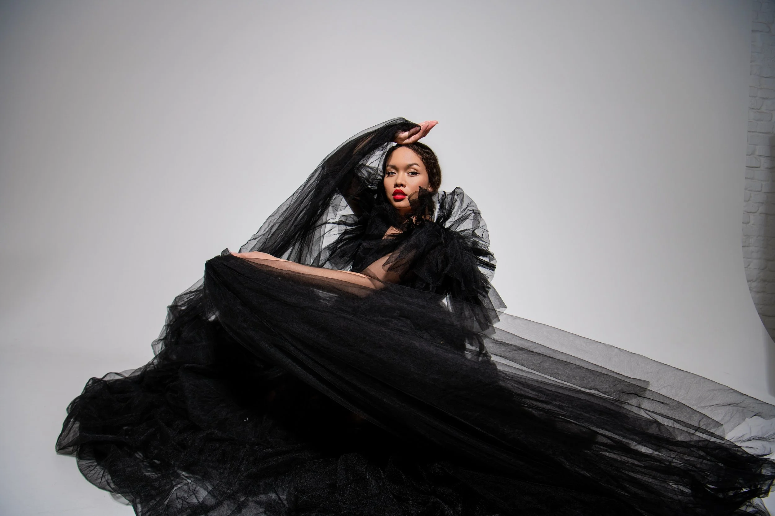 A woman in an elaborate black tulle dress poses against a plain white background, with one arm raised above her head and a confident expression.
