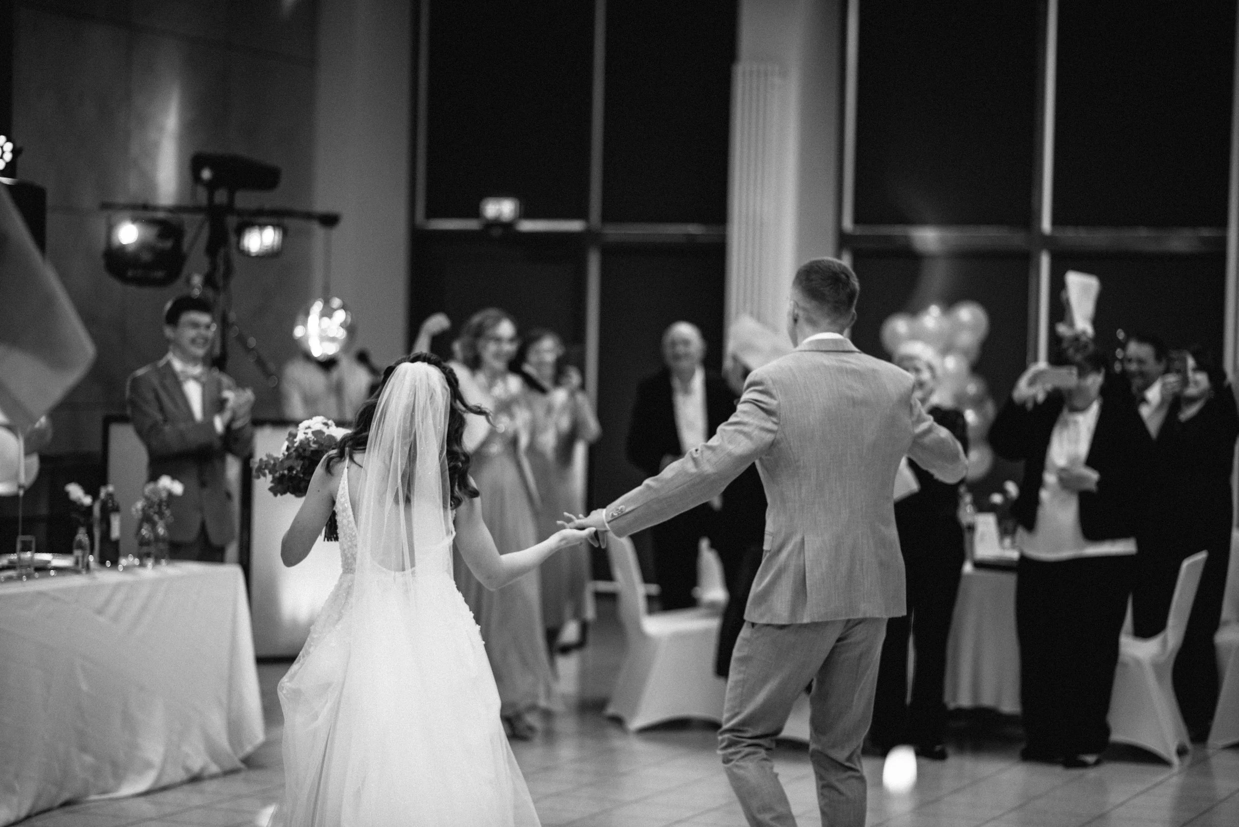 Black and white photo of a bride and groom dancing at their wedding reception, with guests clapping and cheering in the background.