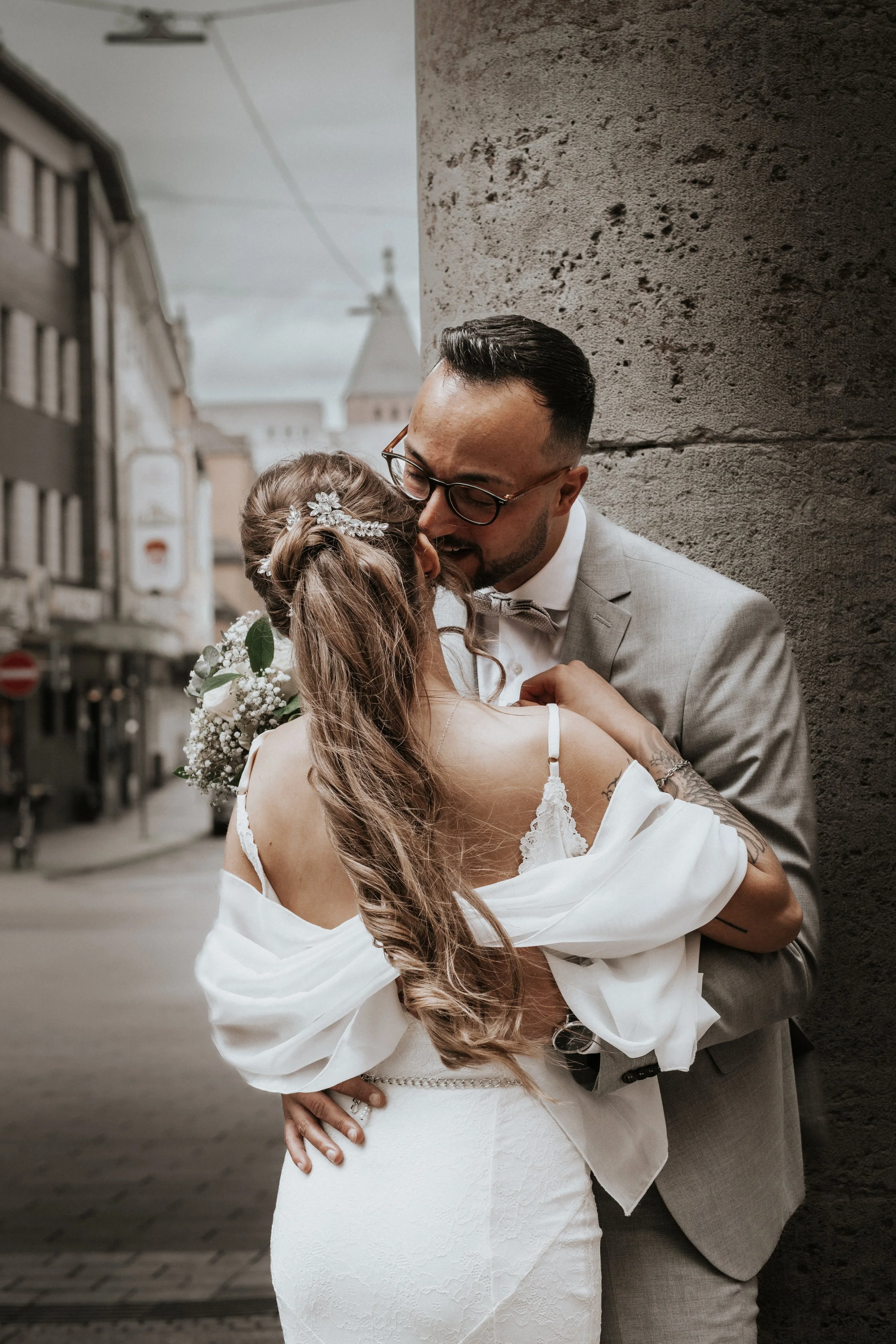 A newlywed couple sharing a kiss on a city street, with the bride in a white wedding dress and the groom in a light gray suit, standing against a textured wall.