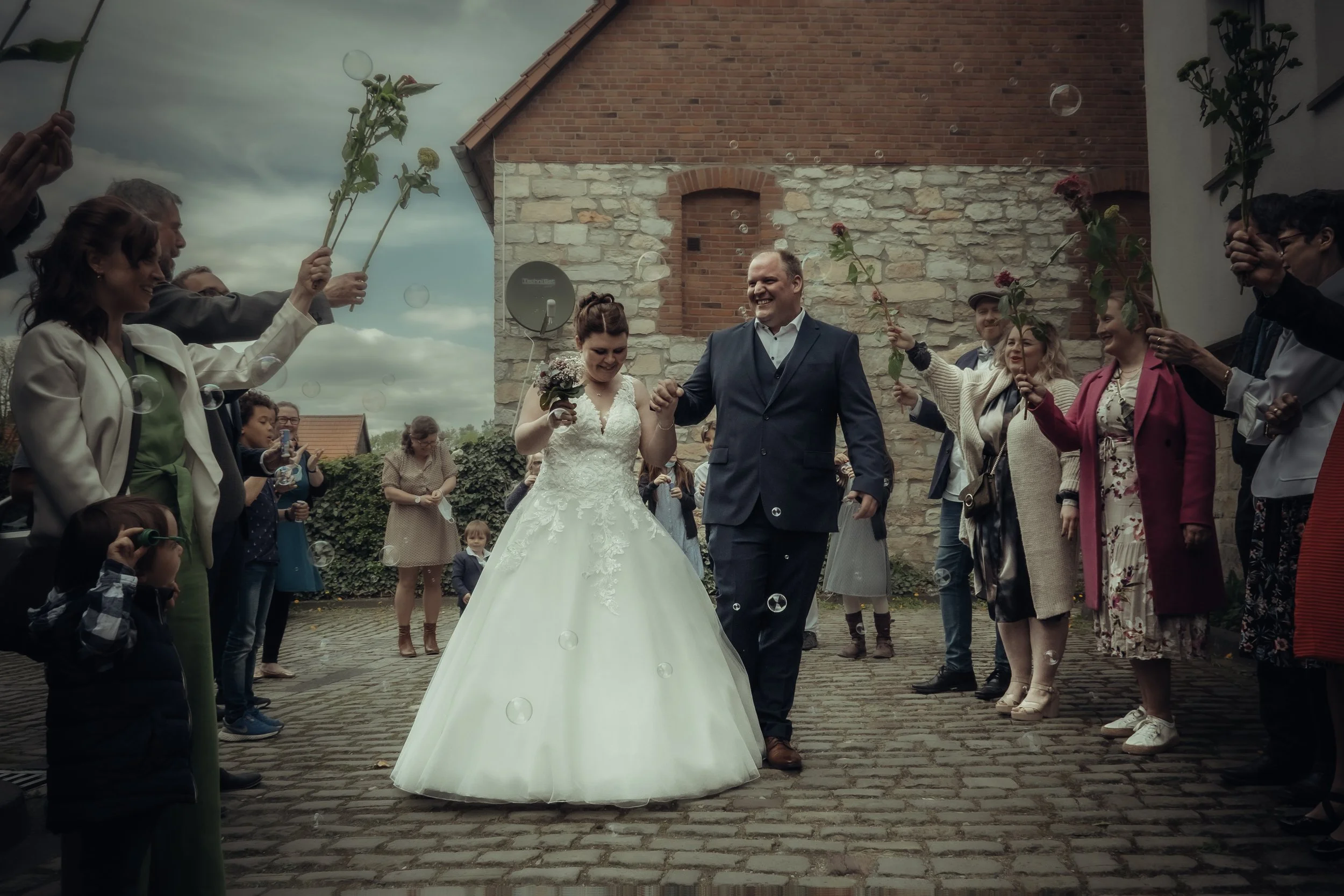 Bride and groom walking hand-in-hand while surrounded by wedding guests blowing bubbles and holding flowers, with a stone and brick building in the background.