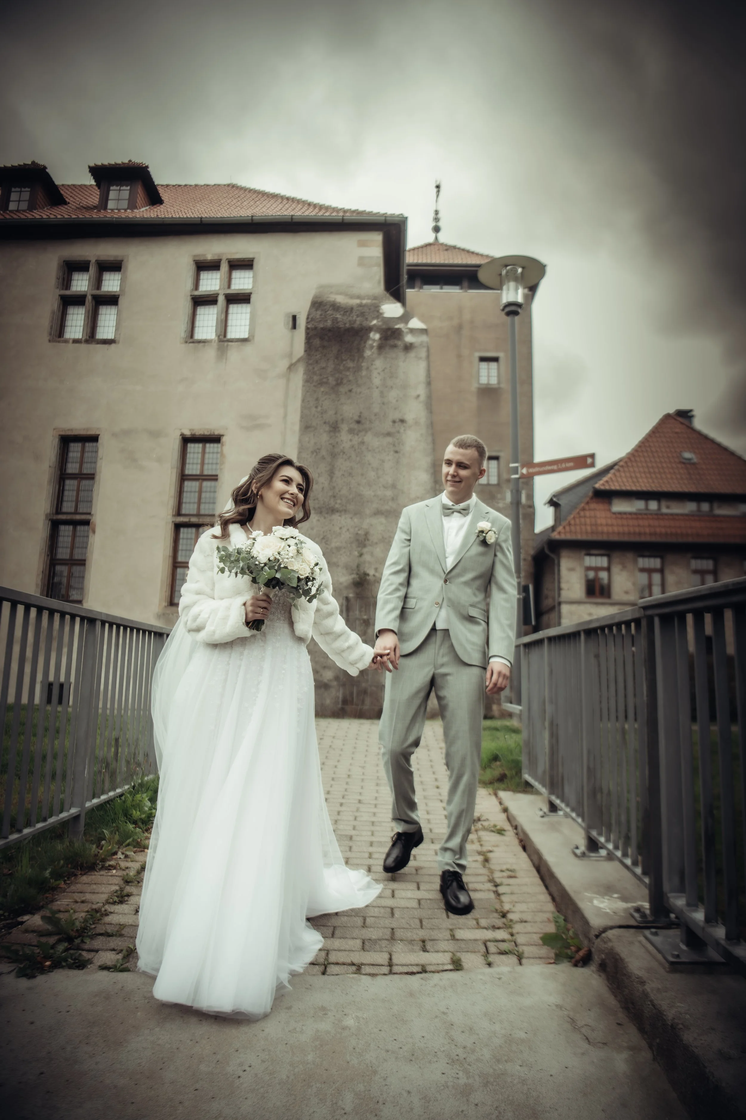 A bride and groom holding hands on a city sidewalk during their wedding day, smiling at each other, with old buildings and overcast sky in the background.