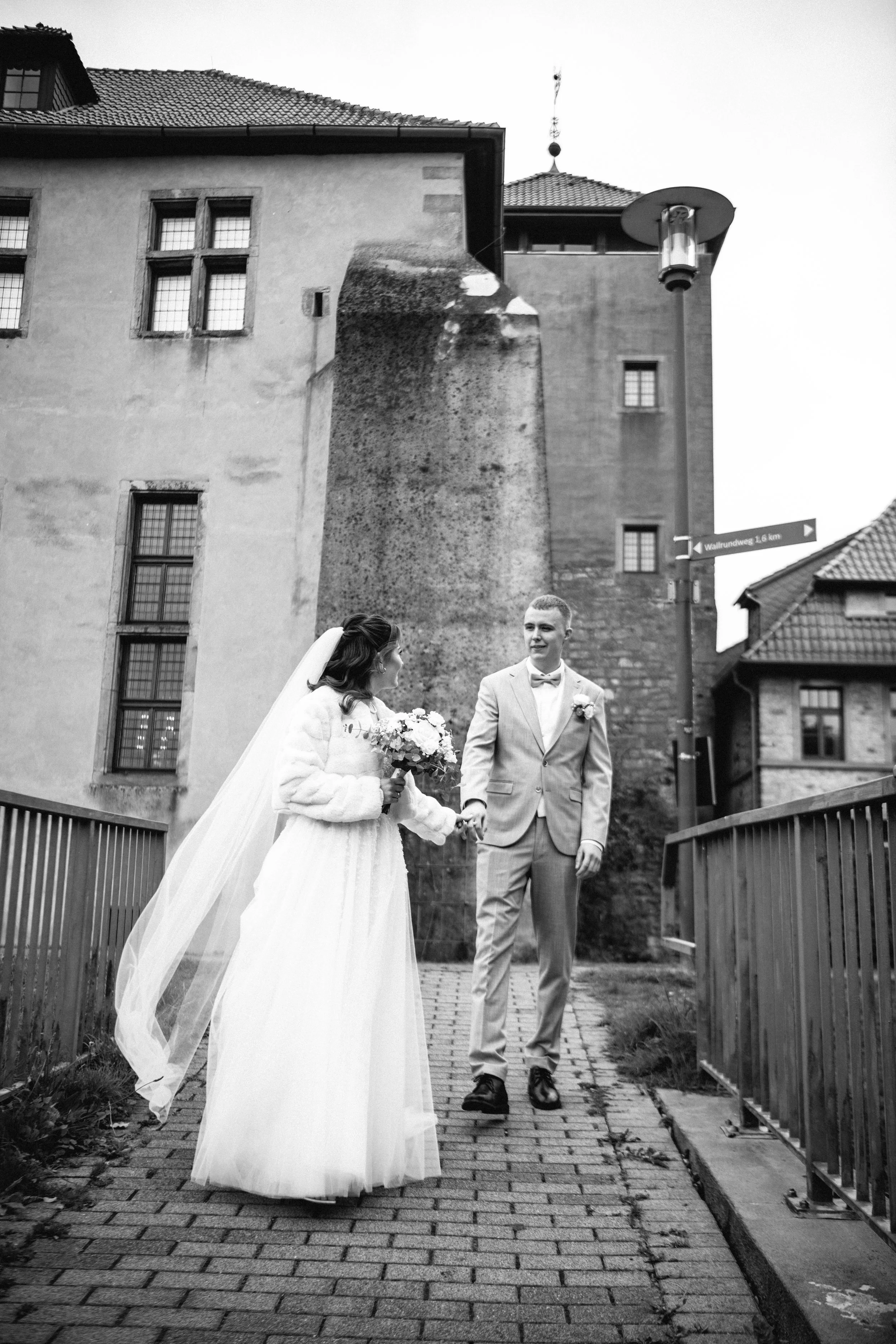 A bride and groom walking hand in hand on a cobblestone path in an urban setting, with the bride holding a bouquet and wearing a long veil, and the groom dressed in a light-colored suit. The background features old buildings and a streetlamp.