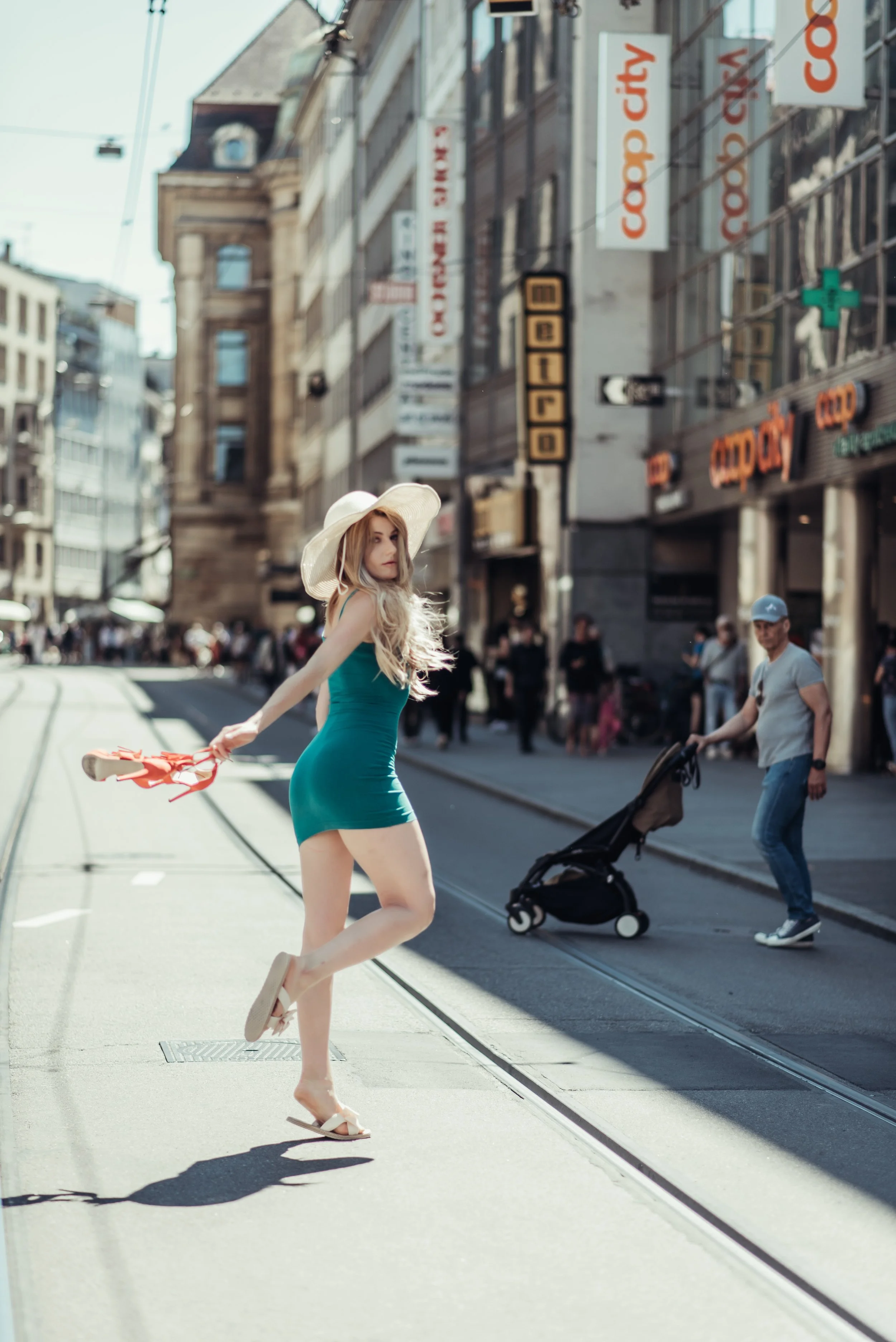 A young woman in a teal dress and wide-brimmed hat stands on a city street holding her shoes, looking to the side. Pedestrians are walking past, and shops and signs line the street.