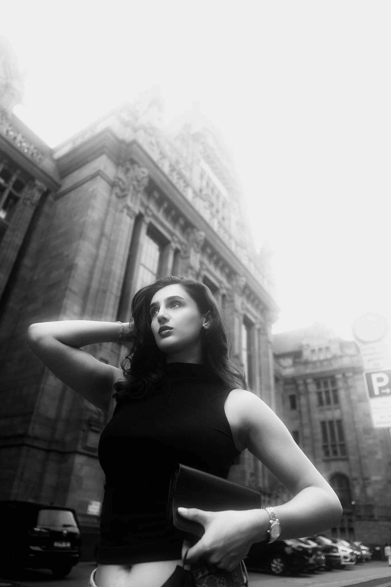 A woman with dark hair wearing a black sleeveless top, standing in an urban environment with historic buildings and parked cars in the background.