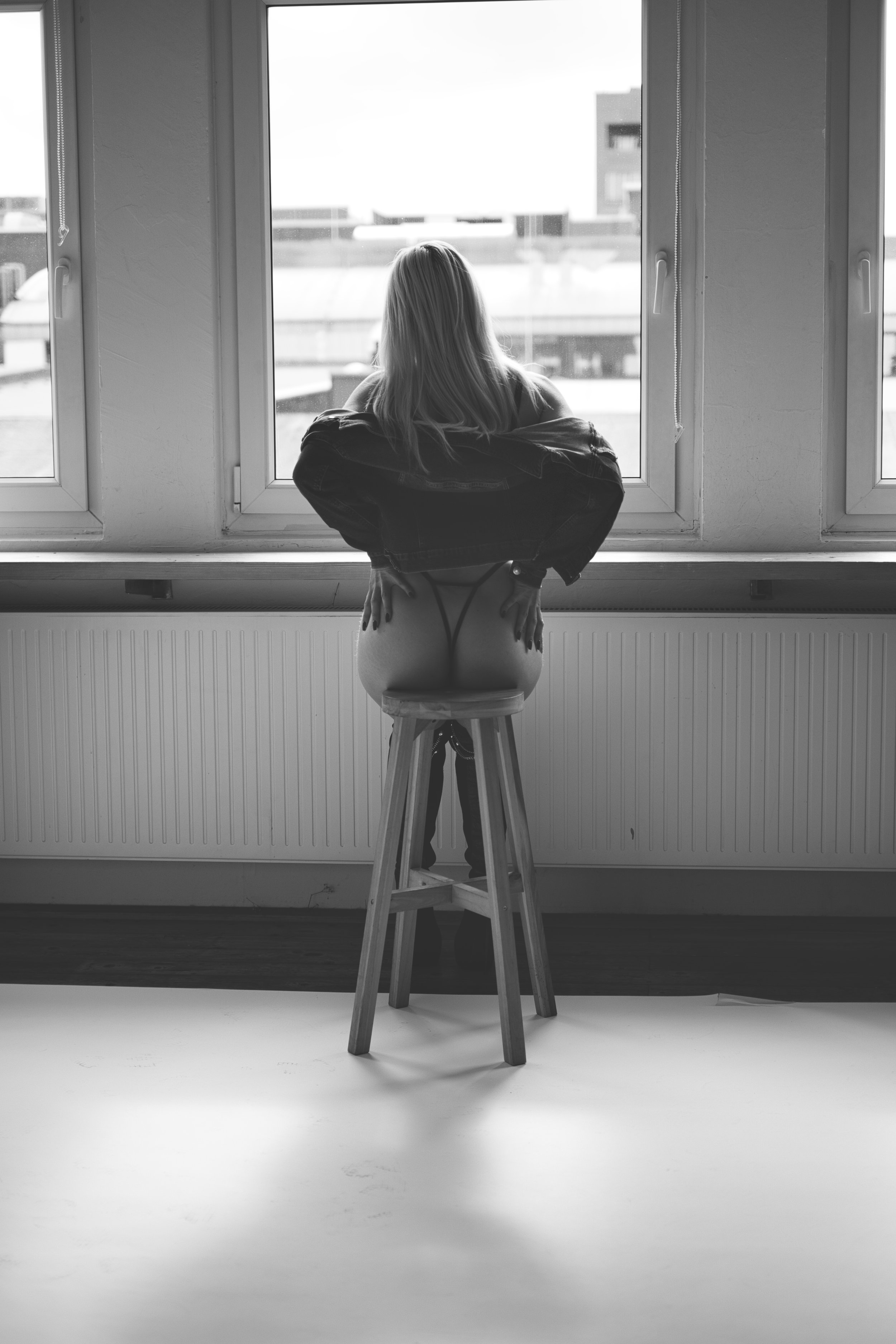 A black and white photo of a woman sitting on a stool in front of a large window, with her back to the camera. She has long hair and appears to be looking out the window, with her arms resting on her knees.