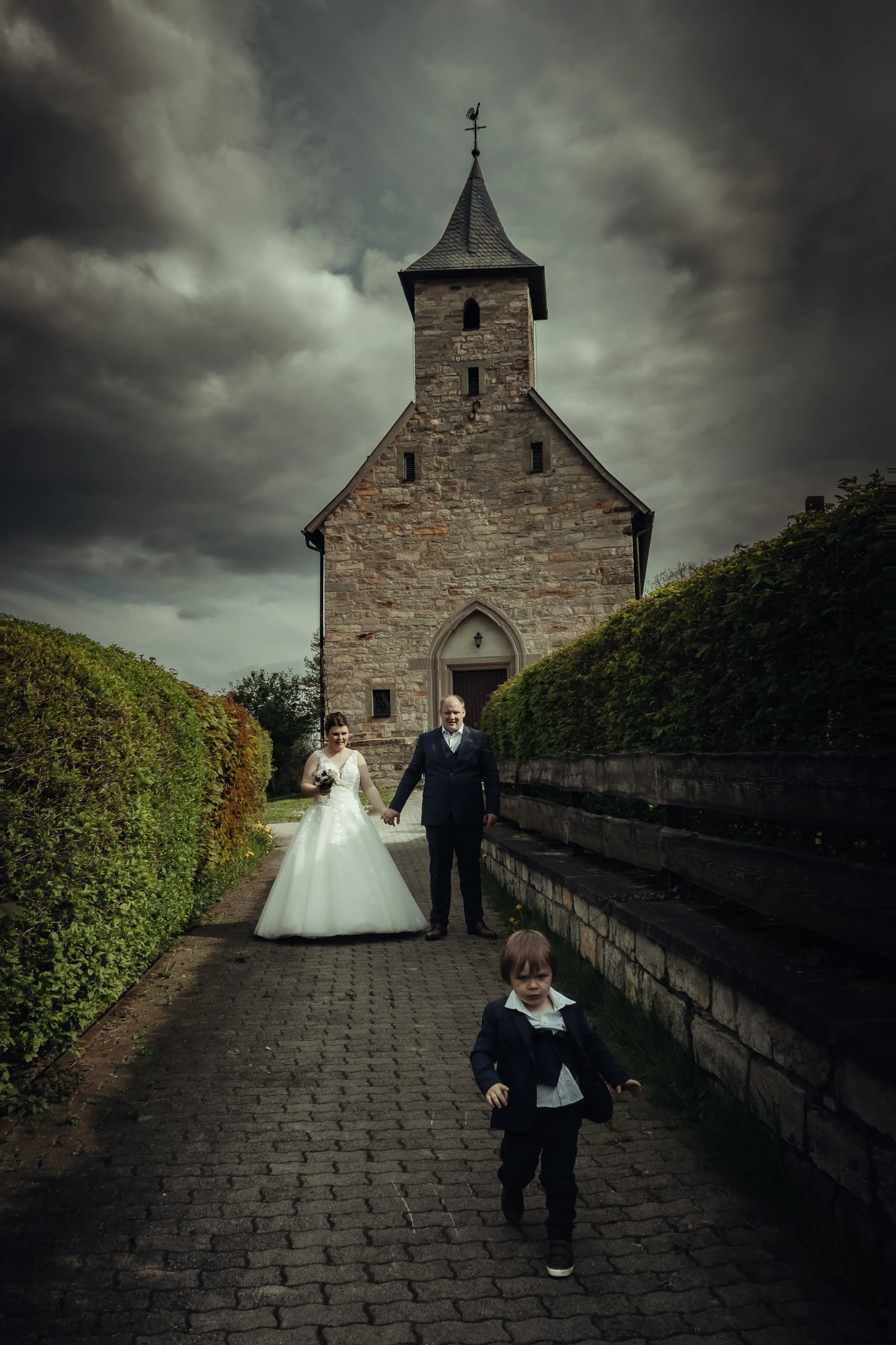 A bride and groom holding hands in front of a stone church, with a young boy walking towards the camera on a cobblestone path, against a dark cloudy sky.