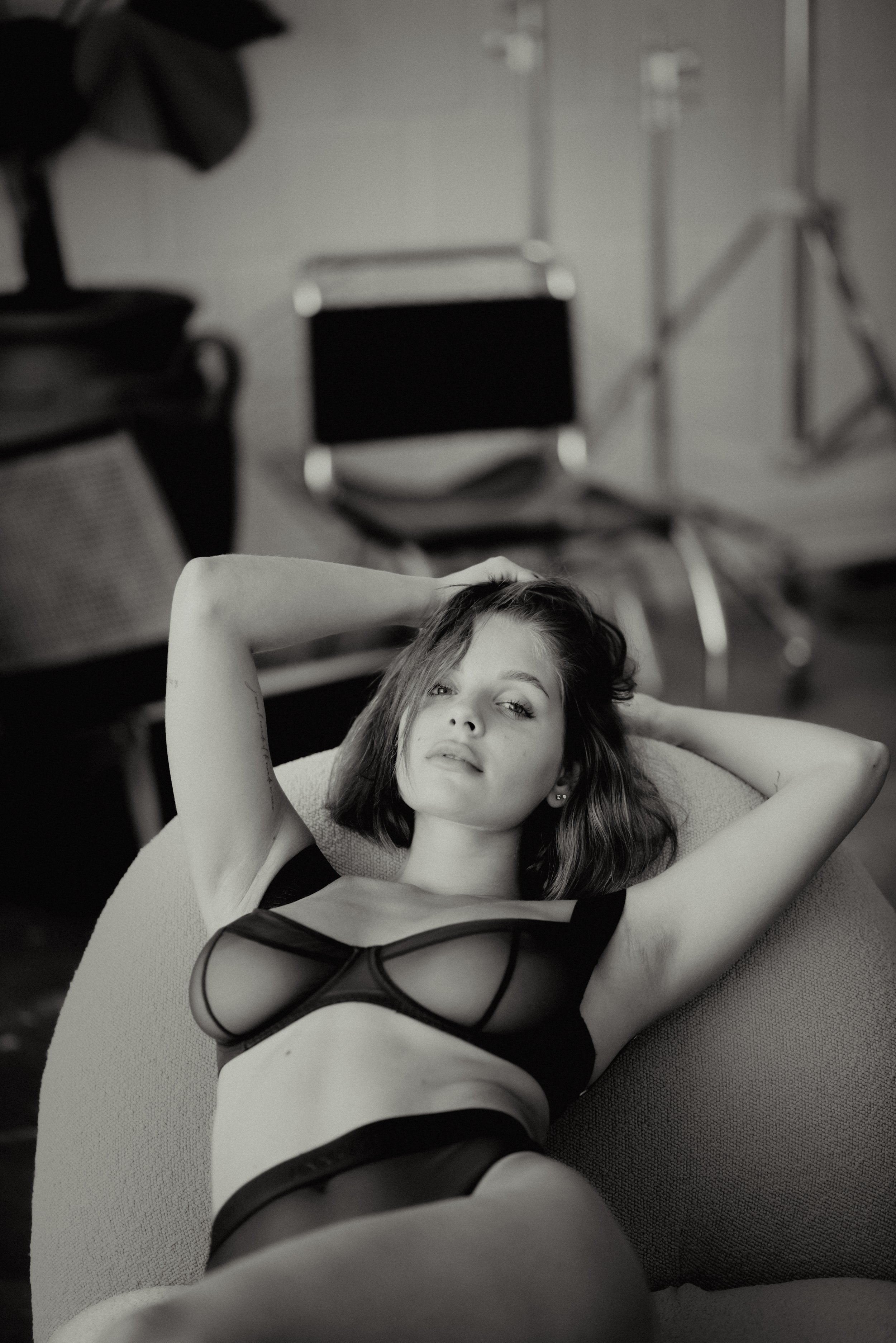 Black and white photo of a woman reclining on a chair with her arms raised behind her head, wearing lingerie, with a blurred background showing chairs and studio equipment