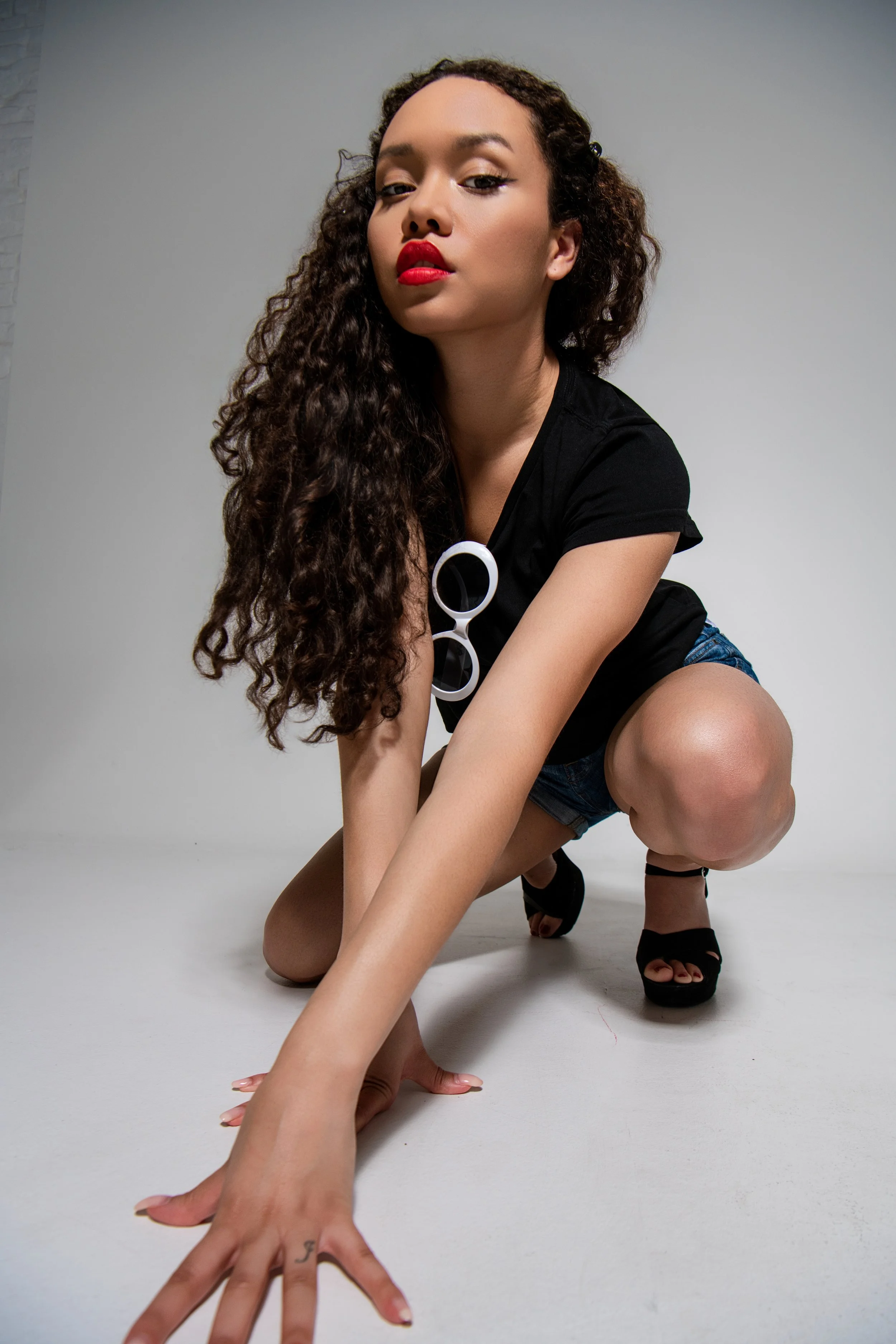 A woman with long curly hair, wearing a black t-shirt and denim shorts, crouching on the floor with one hand extended forward, wearing black high-heeled sandals, and round sunglasses hanging on her shirt. She has bright red lipstick and looks confide